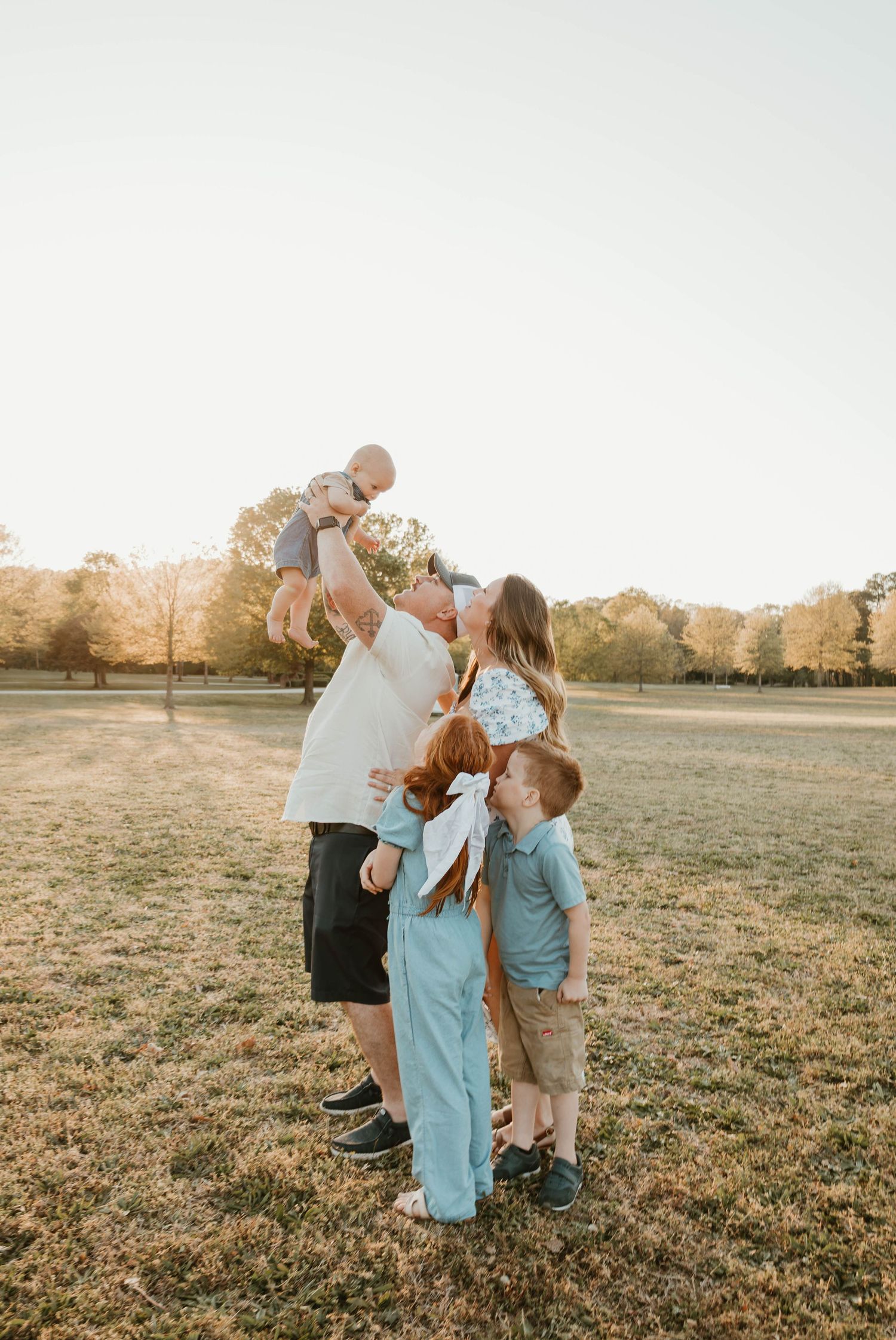 Family of five enjoying golden hour outdoors, dad lifting baby while kids and mom gather close in a sunny park.