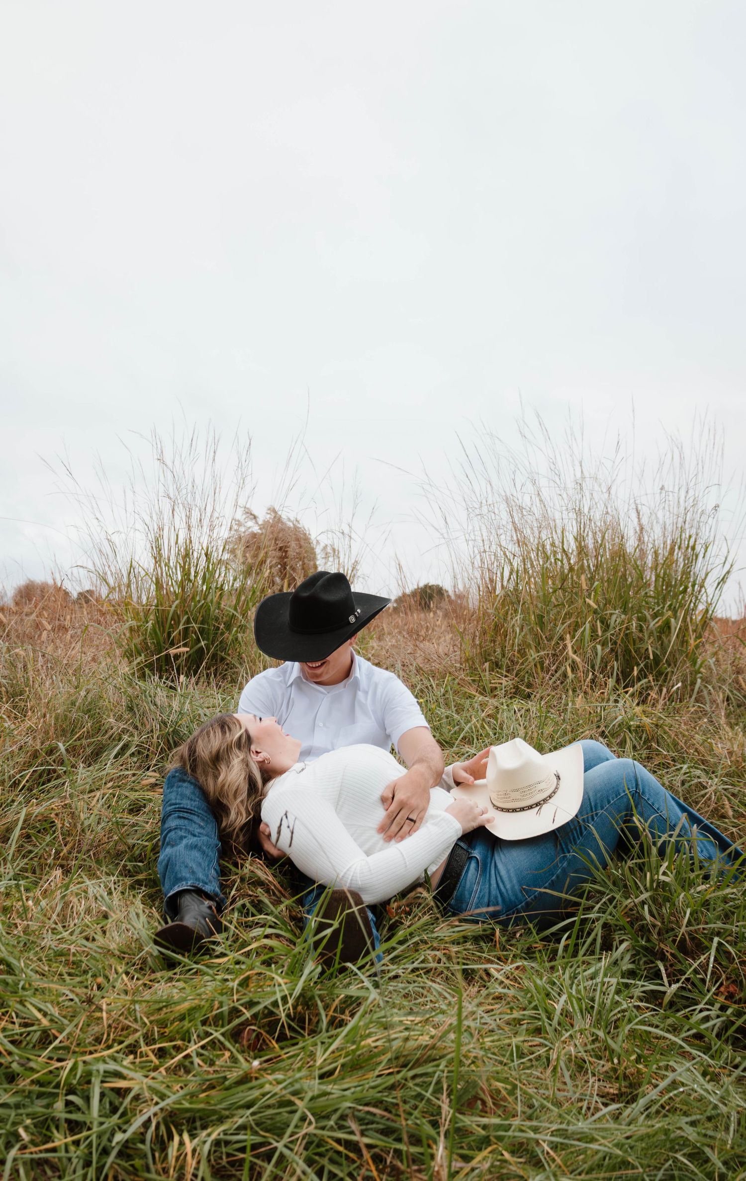Couple lying together in tall grass on an overcast day wearing casual clothes.