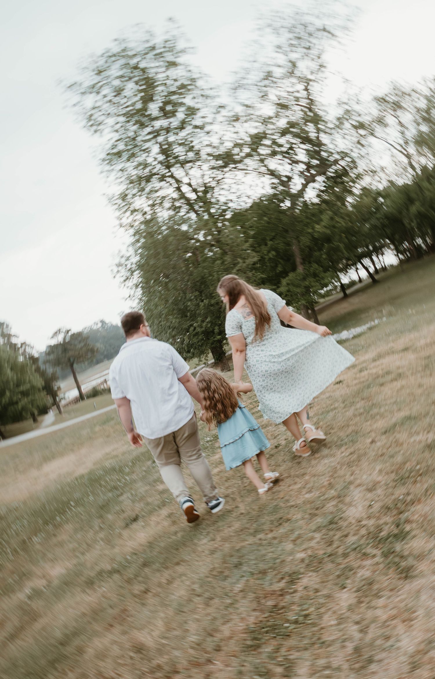 Family of three walking hand-in-hand through a grassy park, captured in motion blur with trees in background.
