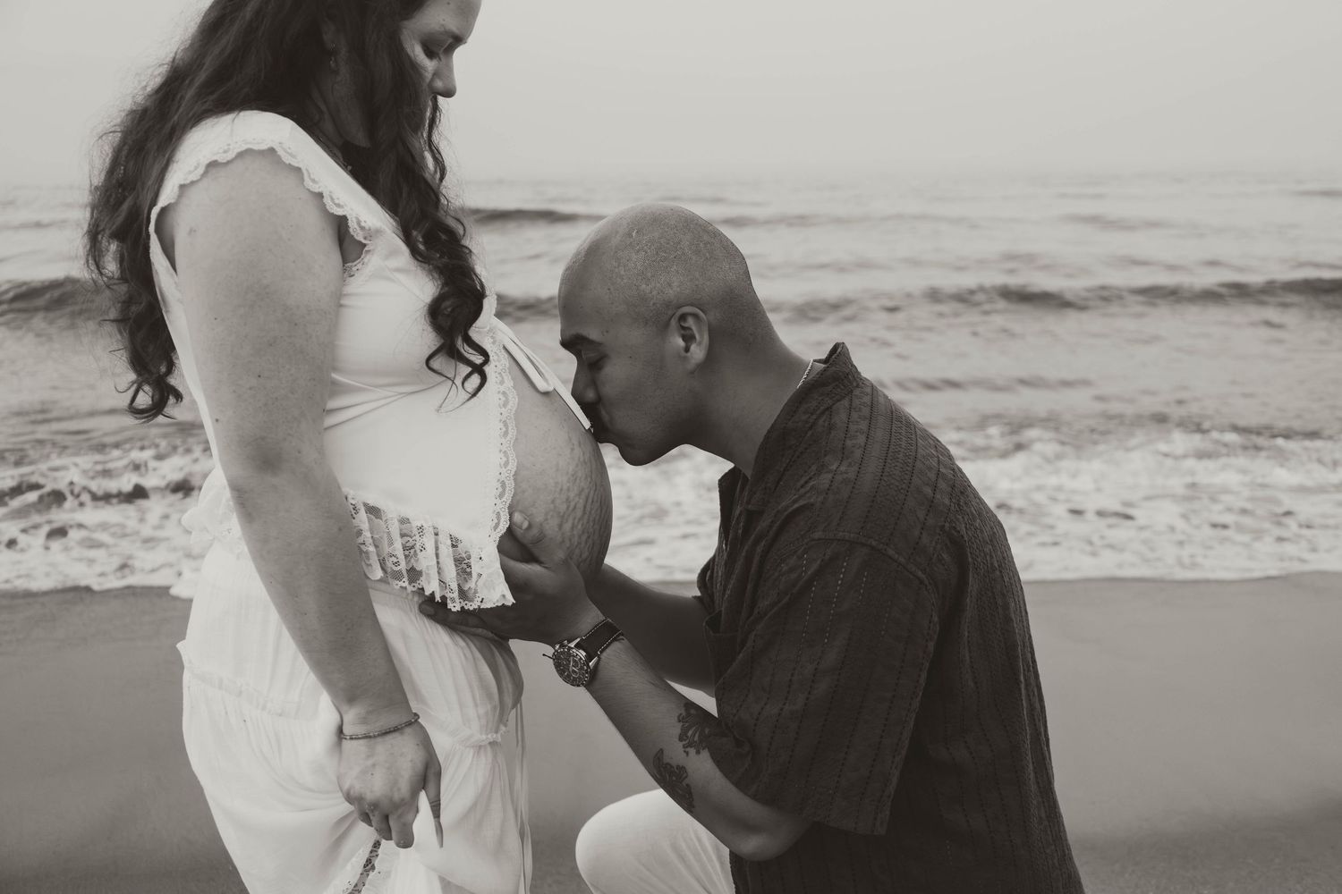 Black and white beach maternity photo of a bald man kneeling and kissing pregnant womans belly by ocean waves.