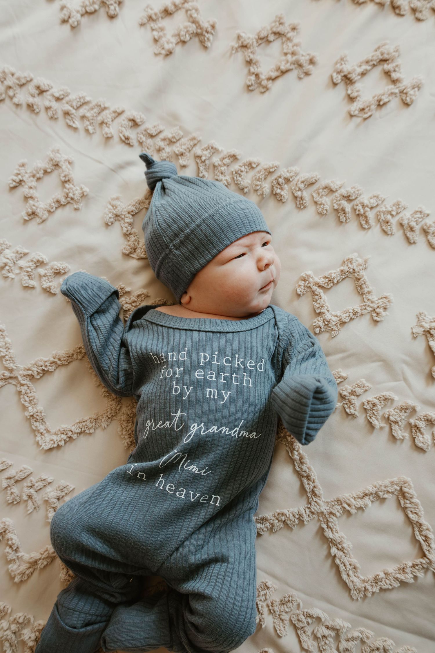 Newborn baby in teal ribbed romper and knotted hat reading hand picked for earth by grandma lying on beige blanket.