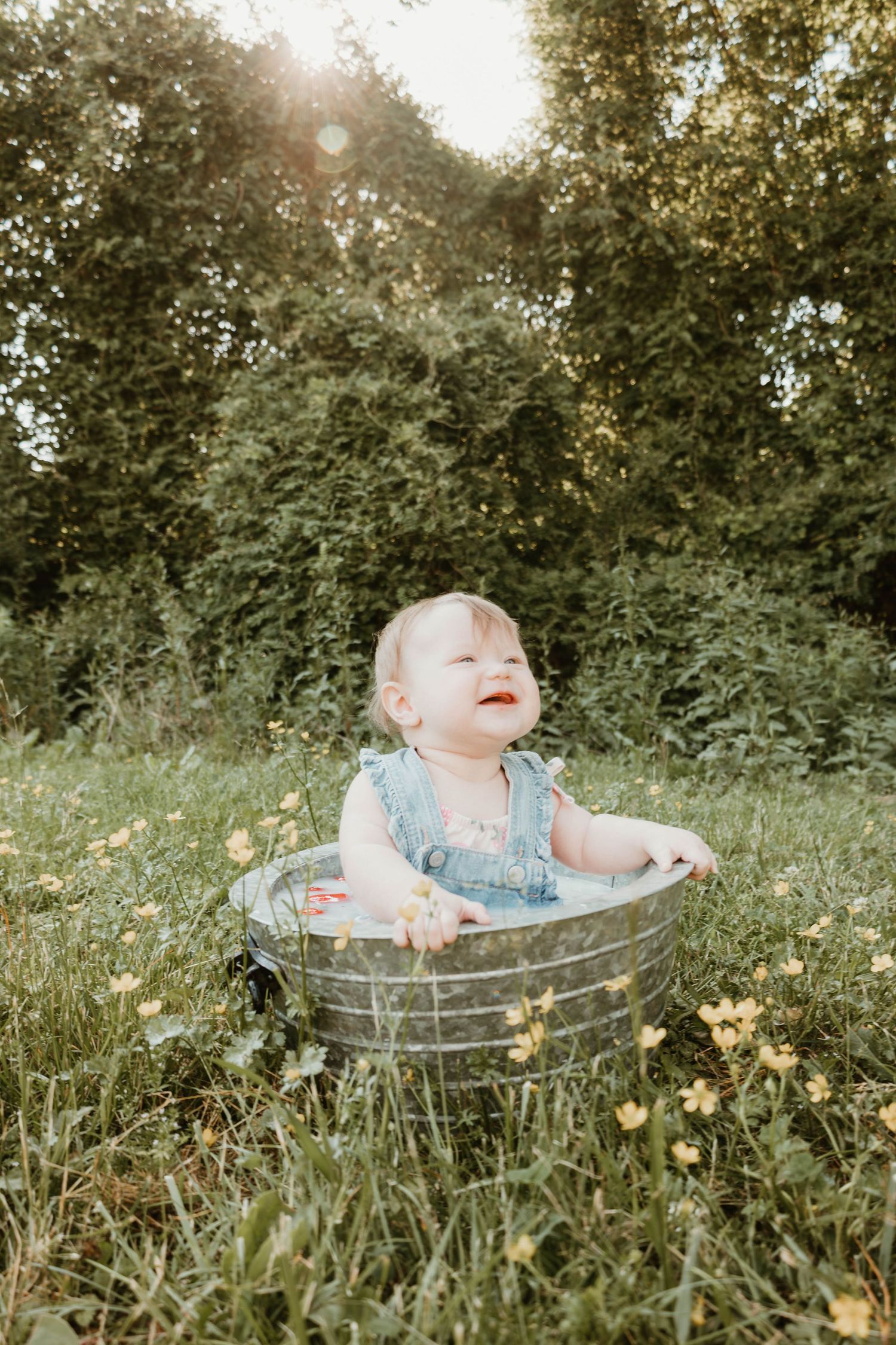 A laughing baby in a denim romper sits in a metal tub outdoors, surrounded by wildflowers and lush green trees.