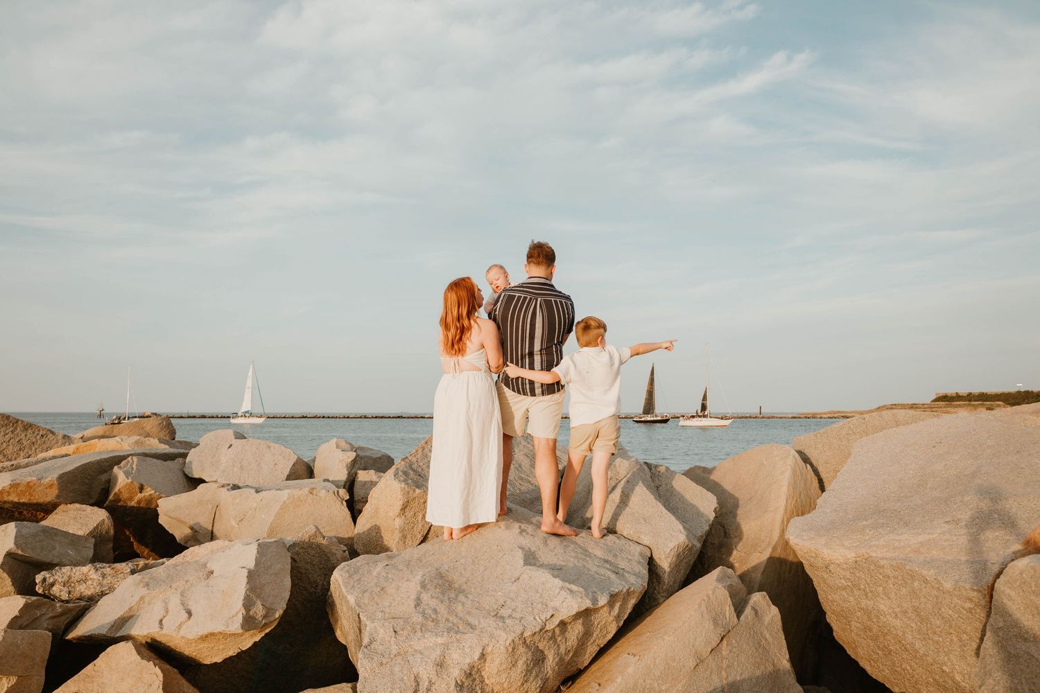 Family of four standing on rocky shore watching sailboats on calm ocean waters at golden hour.