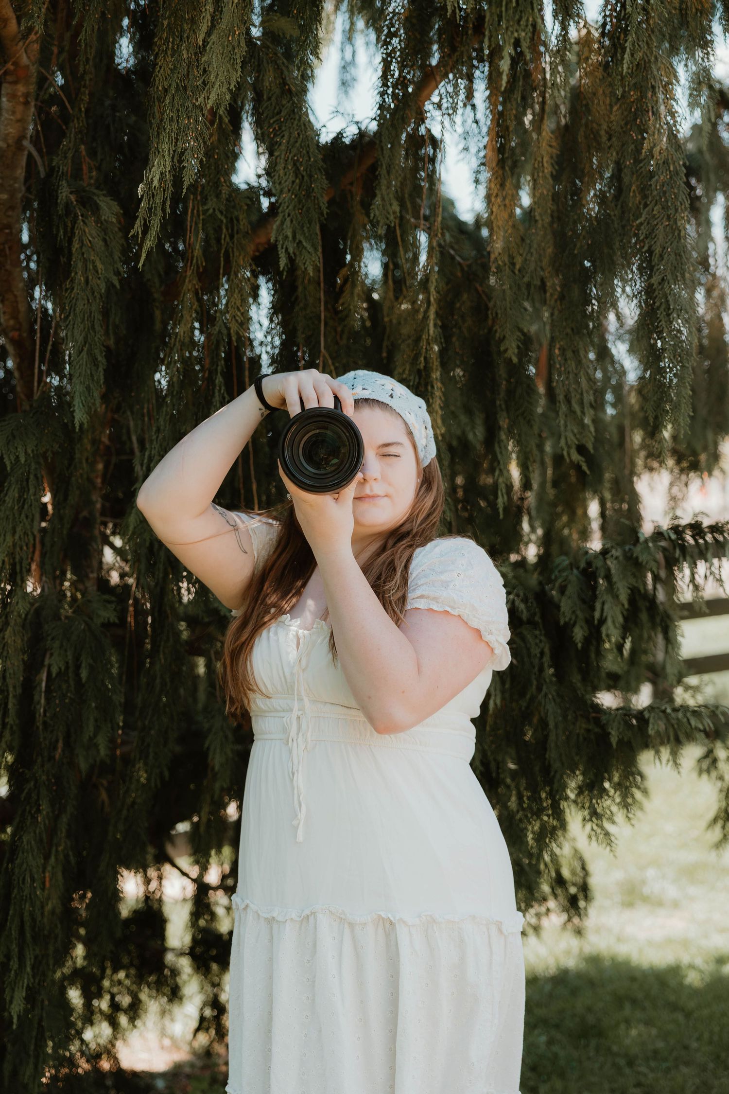 Woman in white dress and headband photographing with DSLR camera outdoors near weeping evergreen tree.