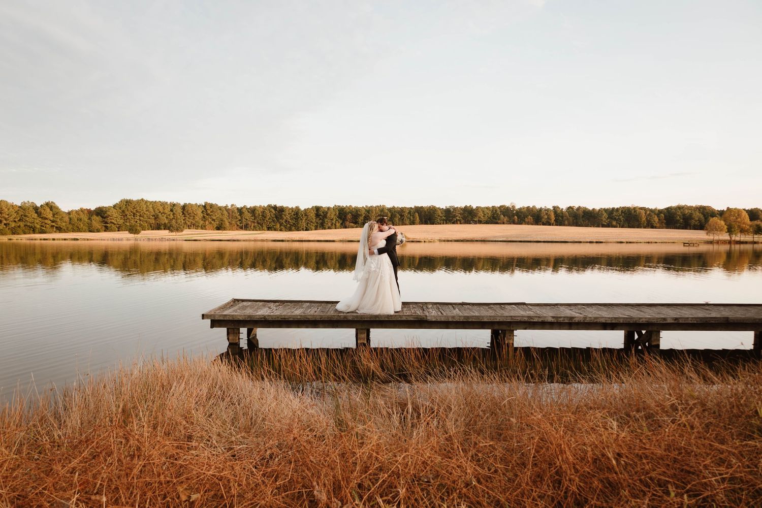 Long wooden dock stretches across a serene lake during sunset at a wedding venue.