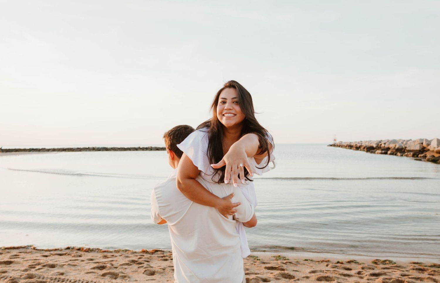 A smiling woman in a white dress being lifted by her partner on a sandy beach near calm water at sunset.