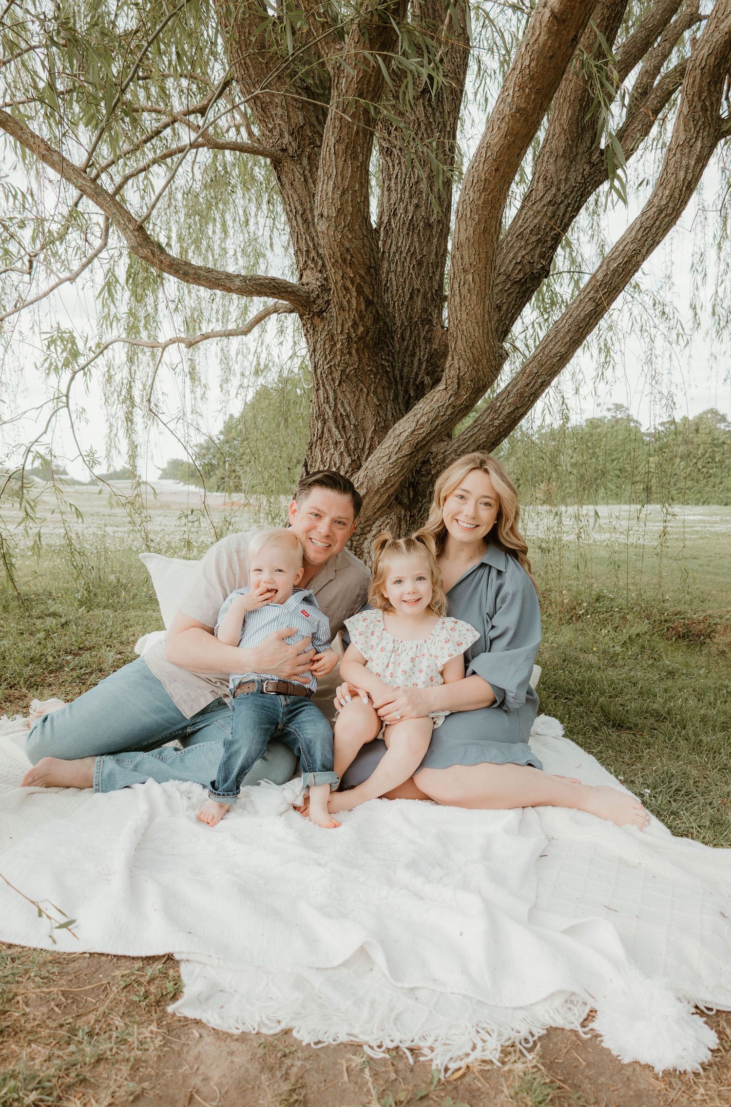 Family of four sitting on white blanket under large willow tree, parents holding young children outdoors.