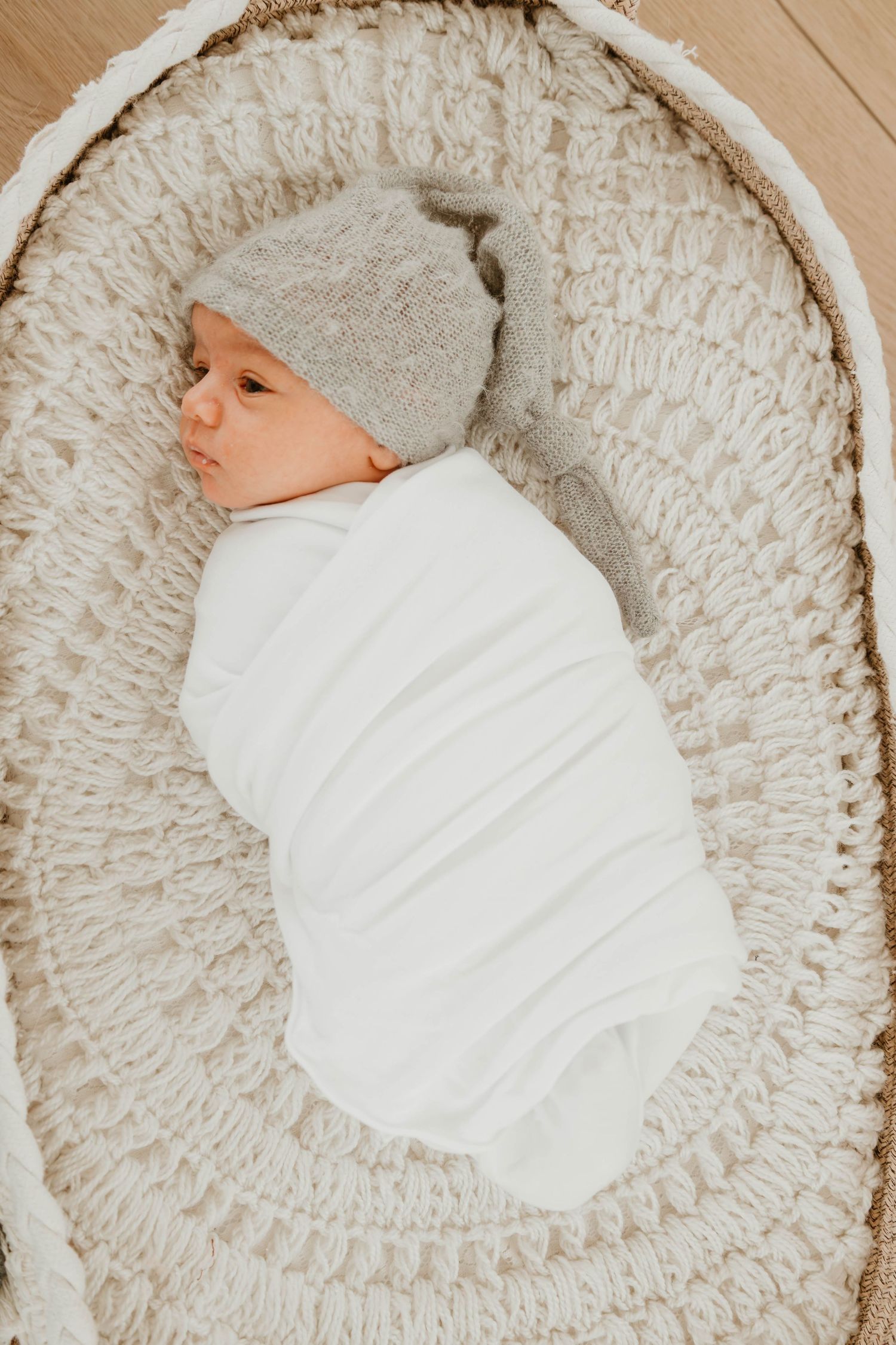 Newborn baby swaddled in white wrap wearing gray knit hat, resting in cream crochet moses basket.