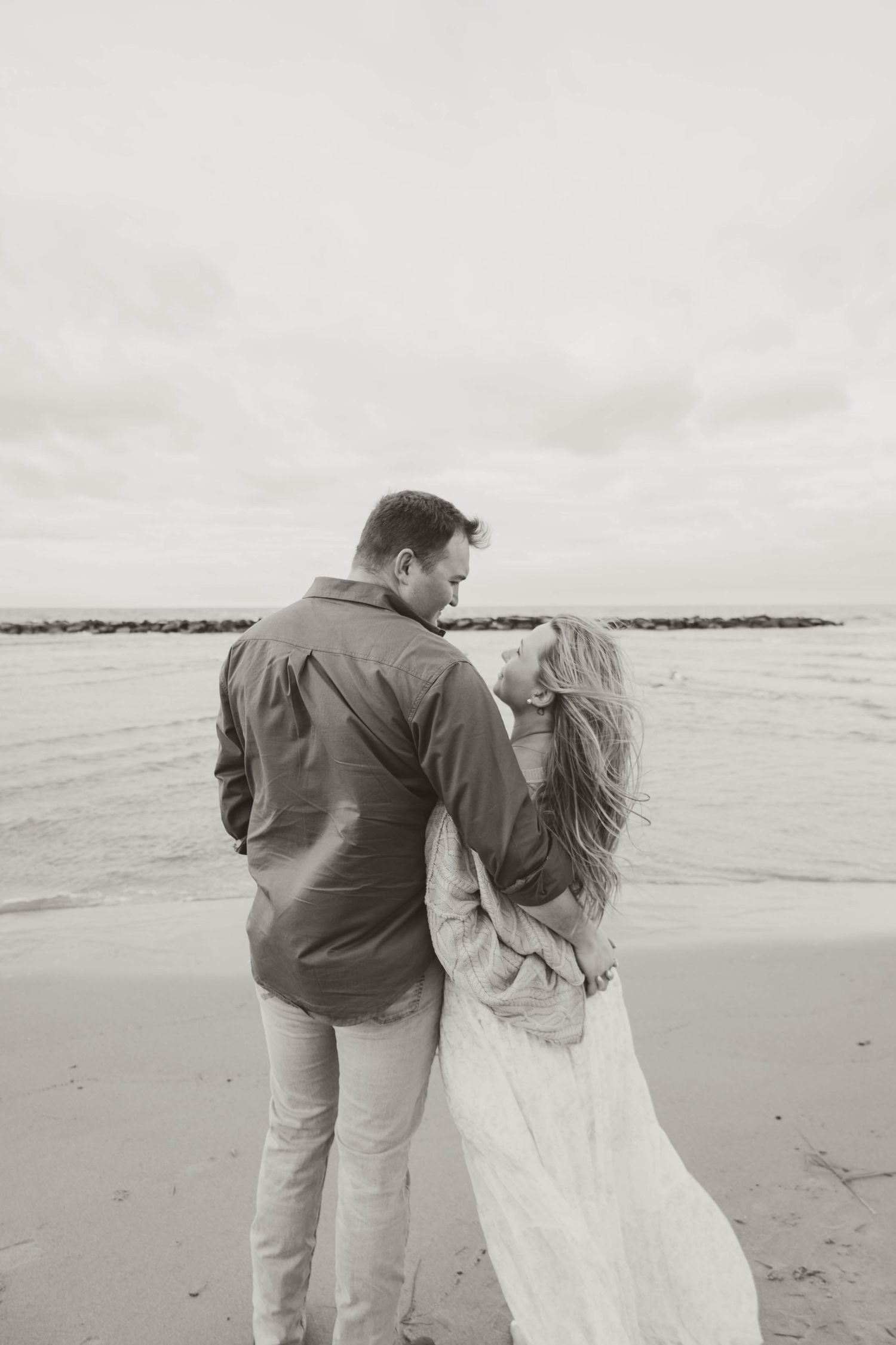 A couple shares a romantic moment walking along a misty beach shore in black and white.