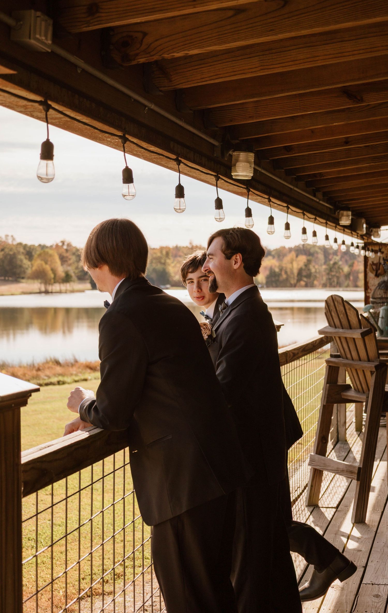 Wedding party gathers on a wooden deck overlooking a serene lake at sunset.