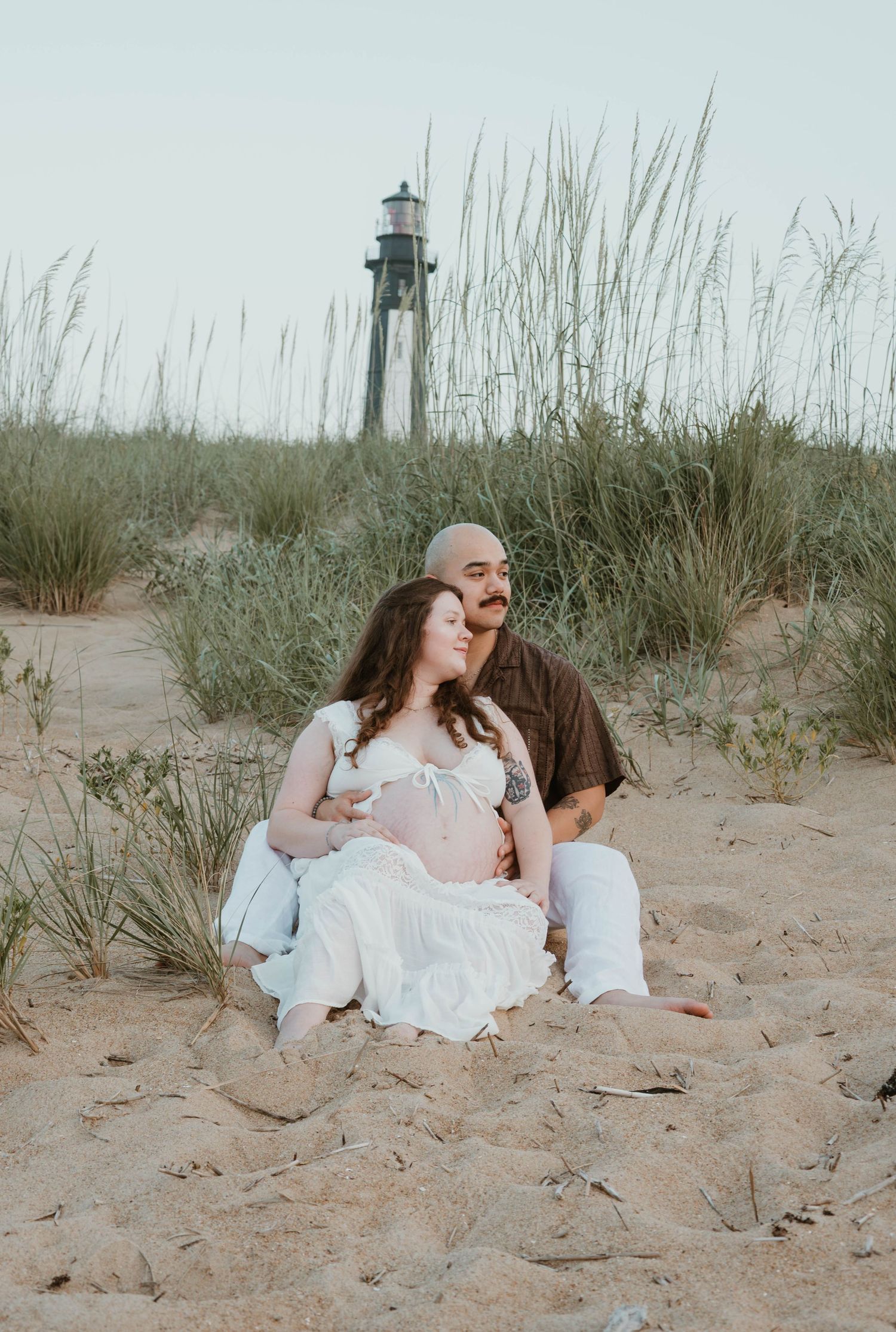 Couple sitting on sandy beach dunes with a lighthouse in background during maternity photo session.