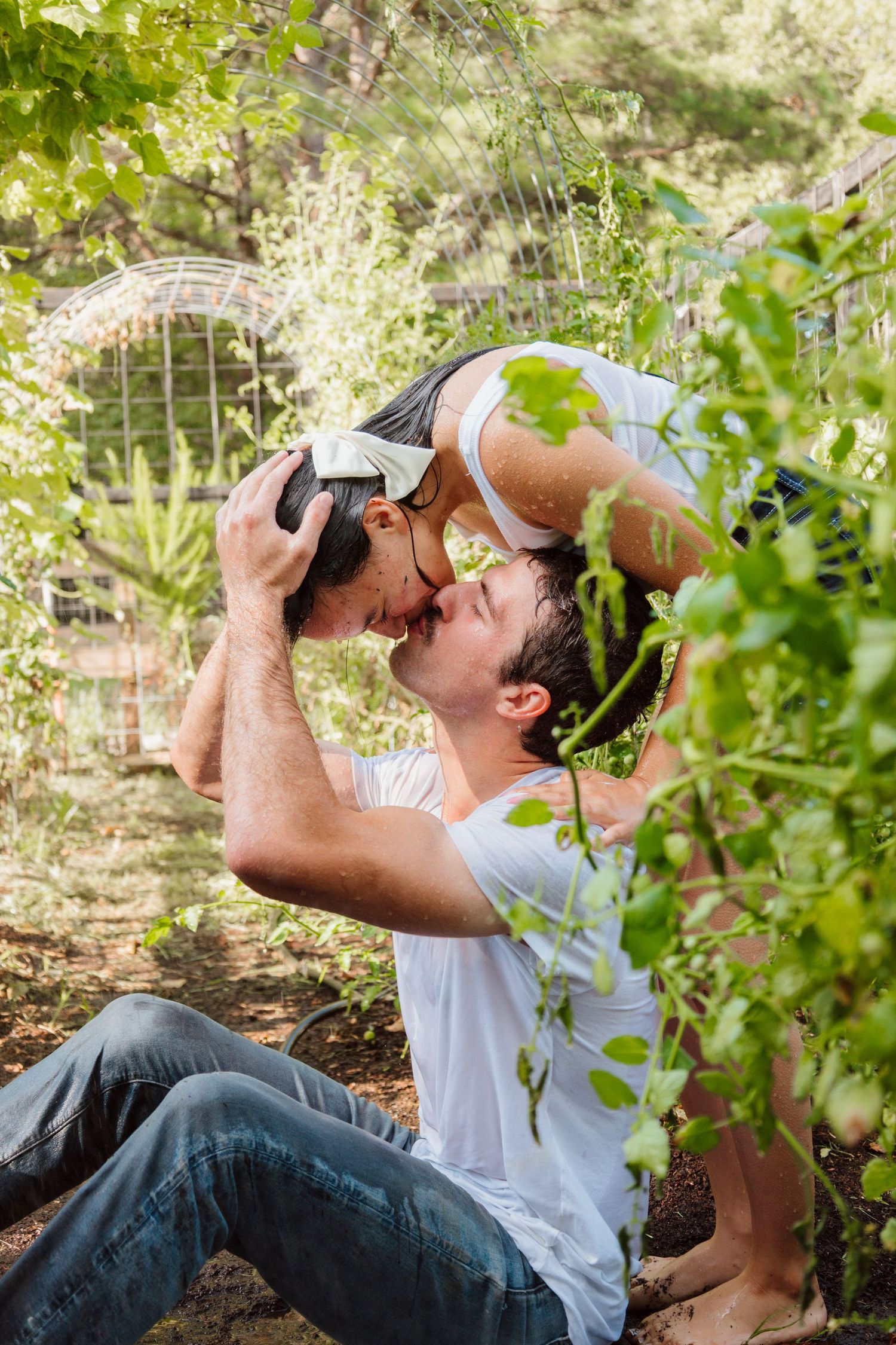 Playful moment captured in a garden as couple shares intimate pose among green foliage.