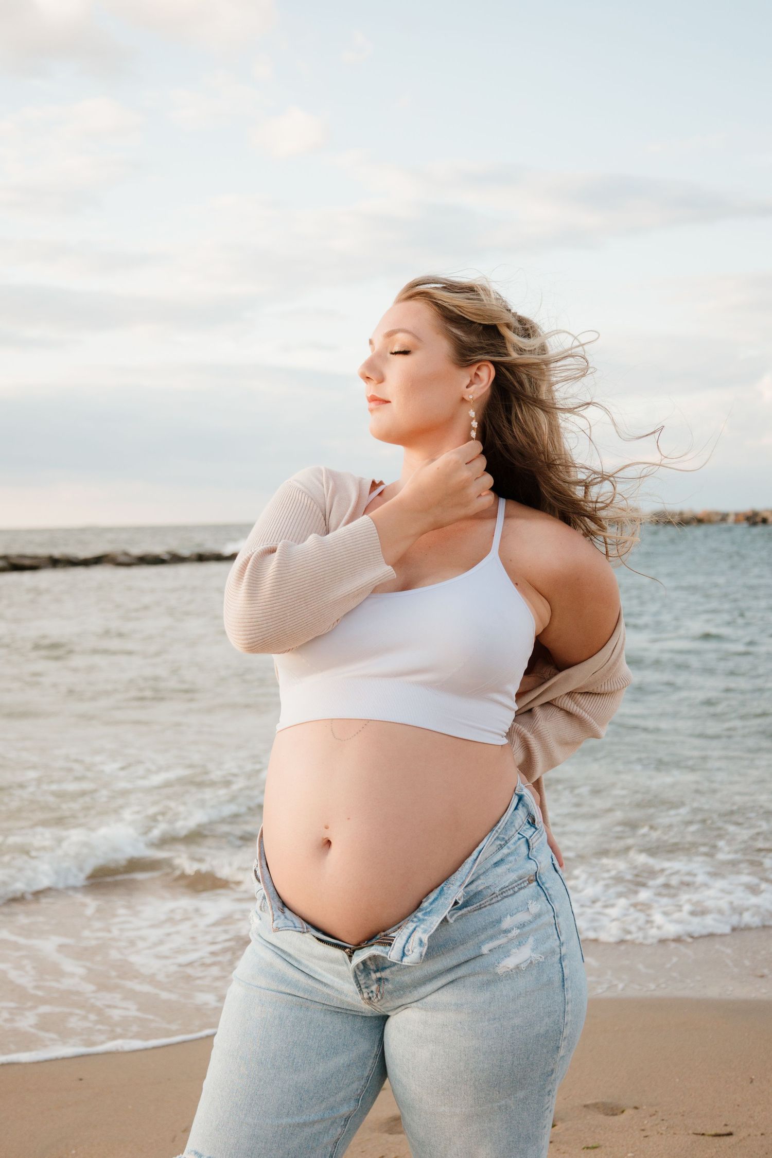 Expectant mother in casual attire poses on a beach at sunset with wind blowing through her hair.