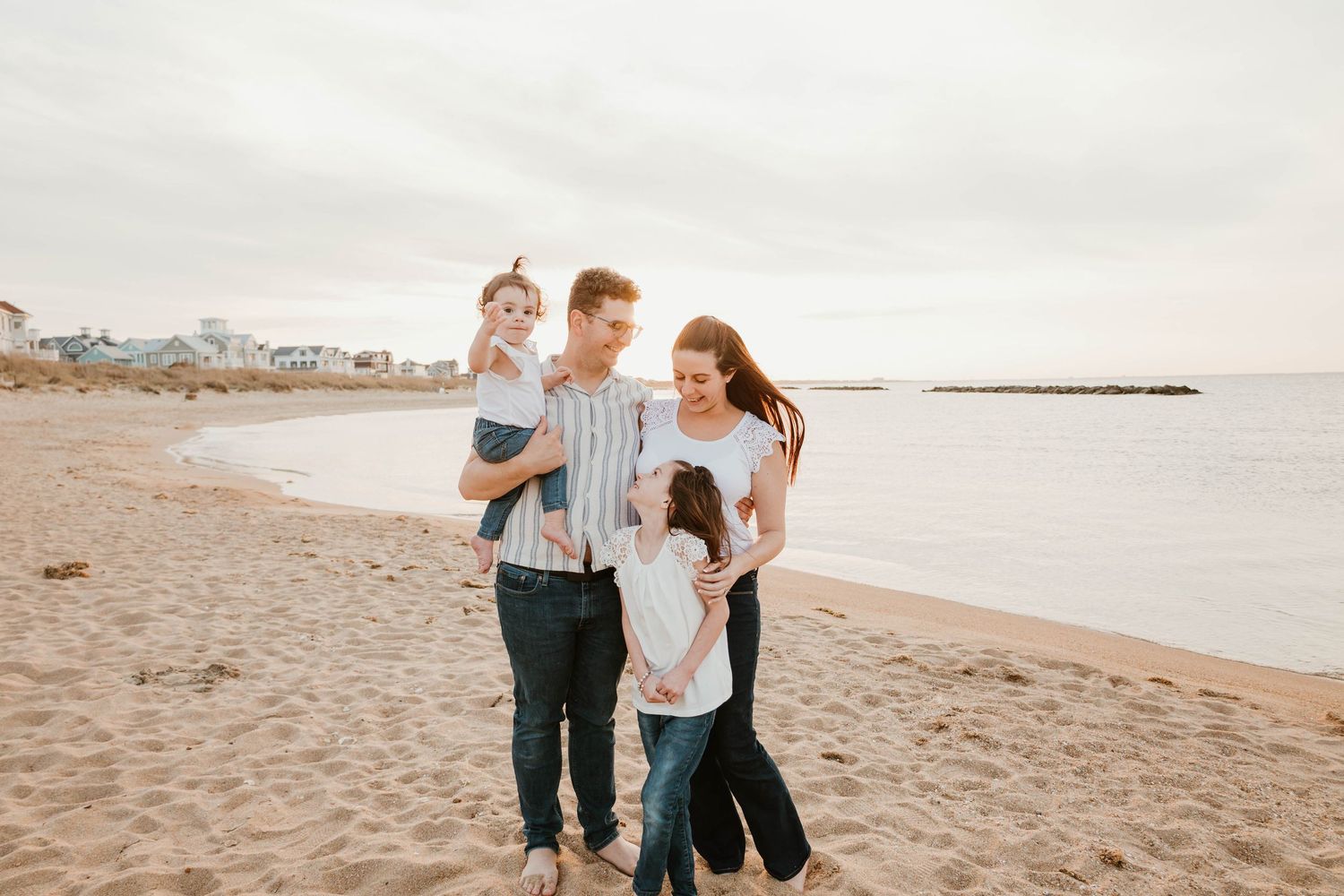Family of four walking on a sandy beach near the ocean, smiling and enjoying a casual outdoor day together.
