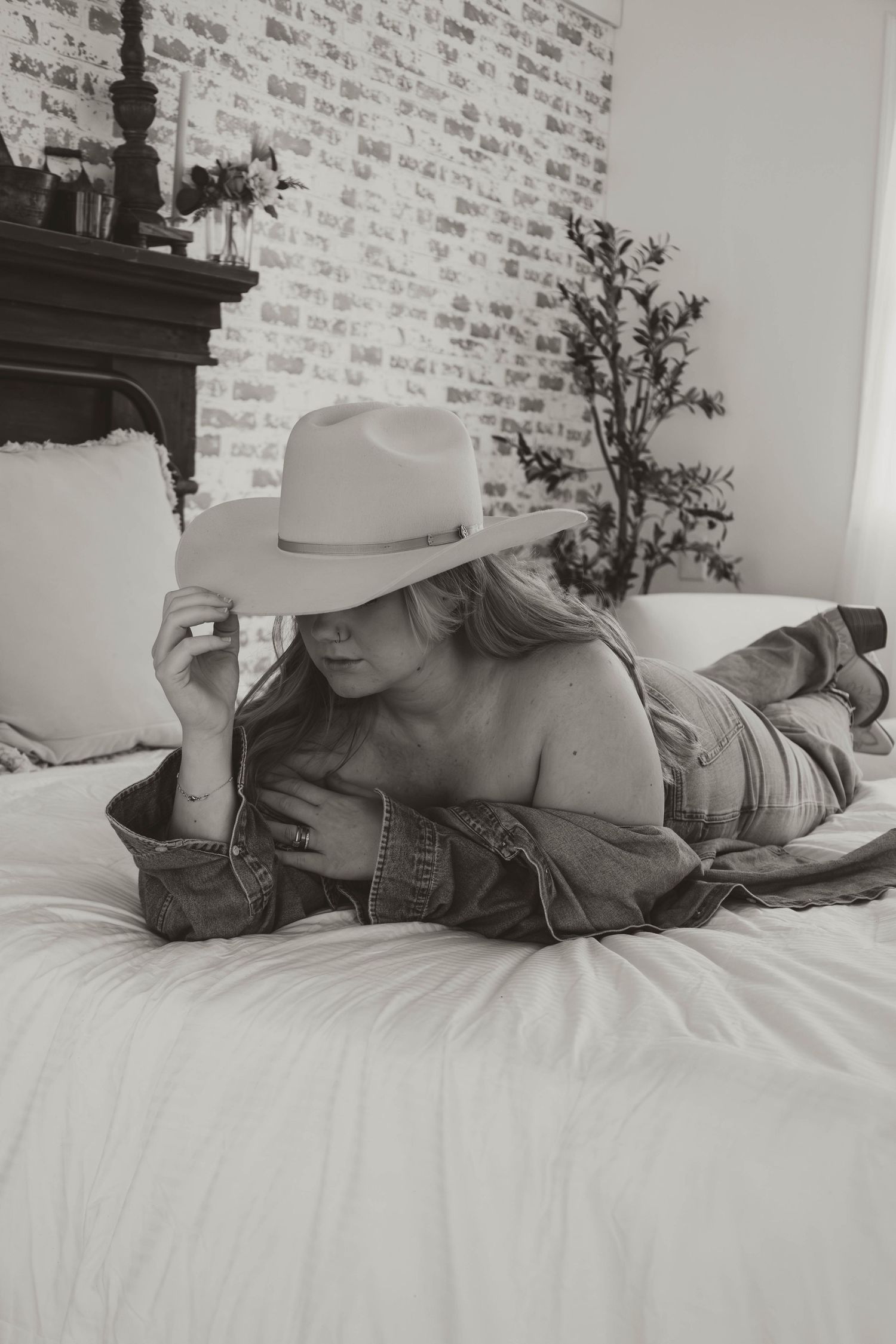Woman in cowboy hat and denim jacket lying on white bed in black and white photo with brick wall background.