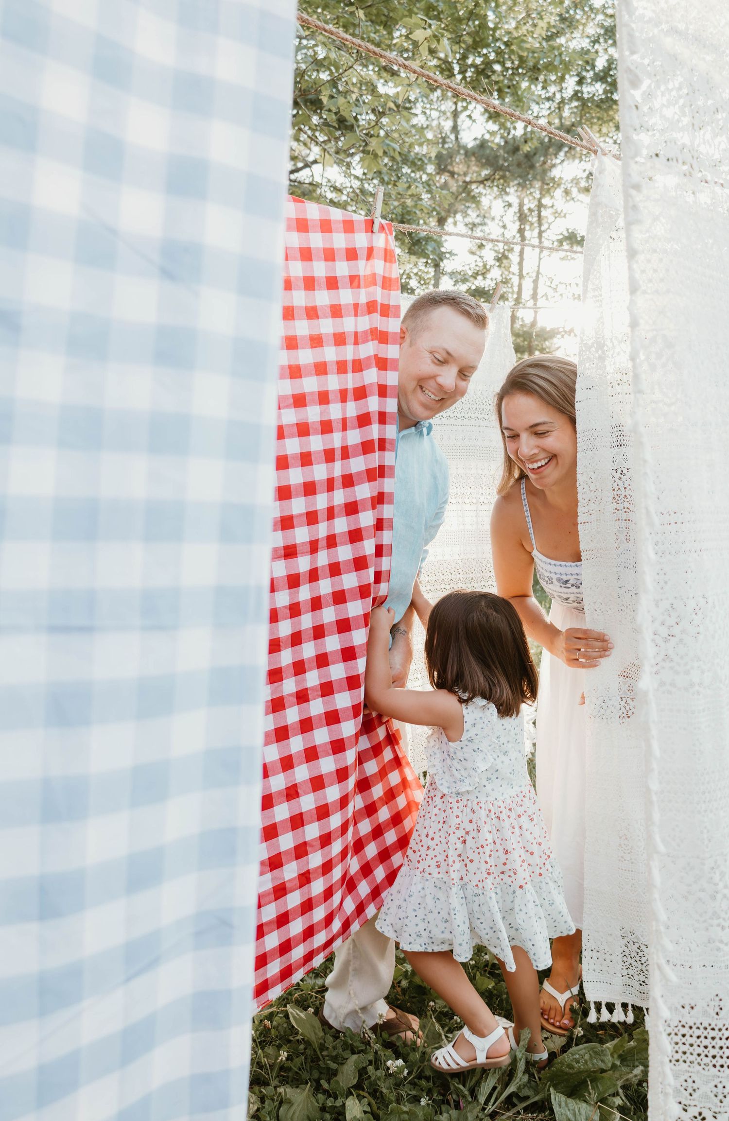 Family of three laughing and playing among hanging checkered picnic blankets outdoors on a sunny day.