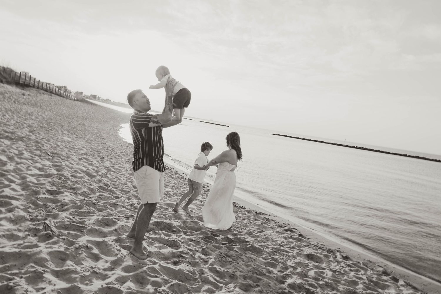 Black and white photo of a joyful family playing on a sandy beach, parents lifting baby in the air near the ocean.
