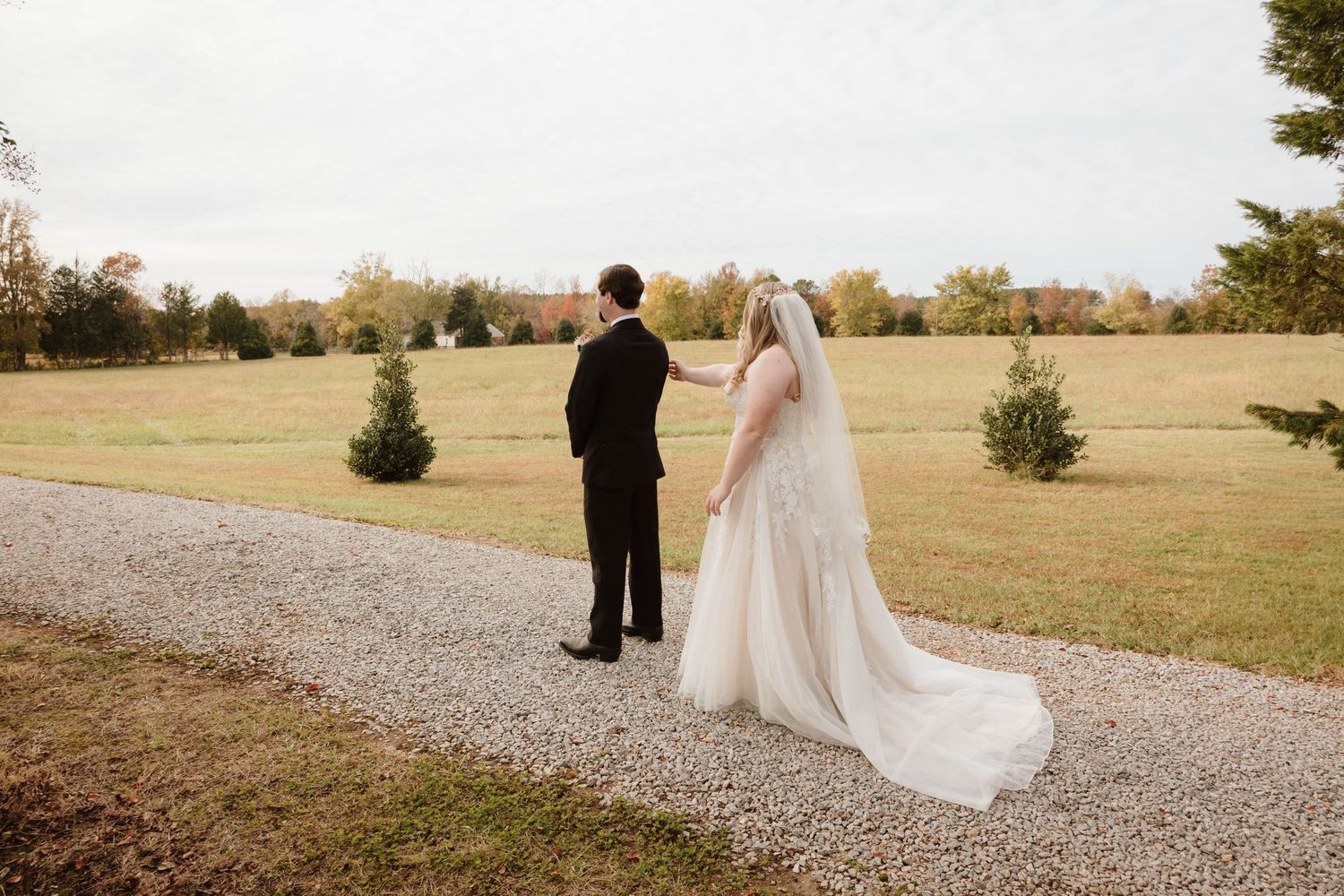 Newlyweds walking along a gravel path surrounded by autumn trees and open fields.