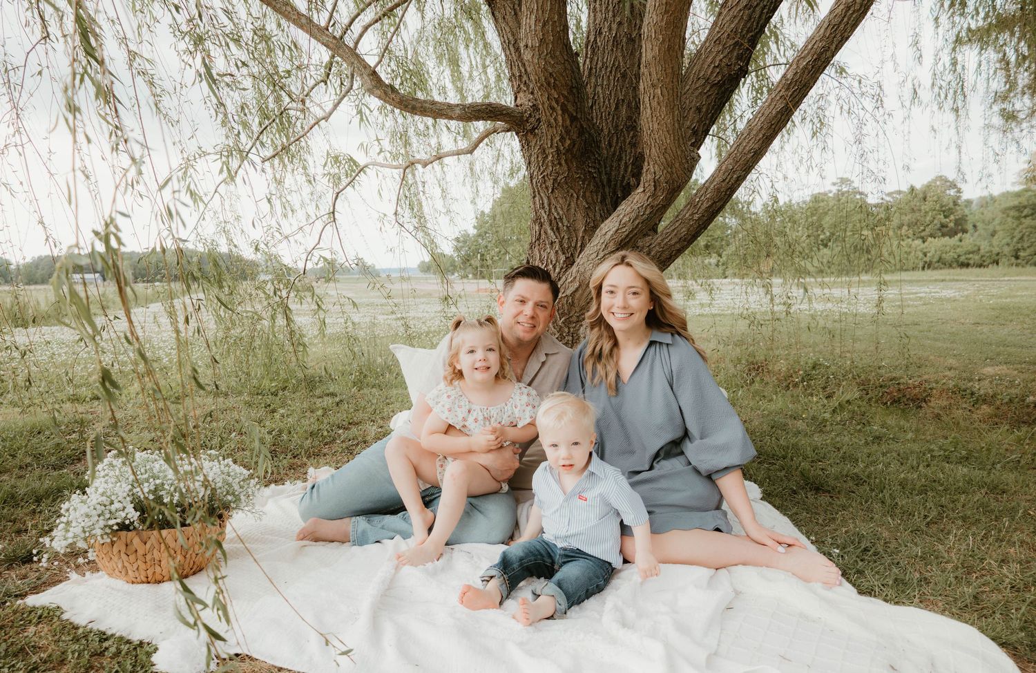 Family of four posing under a willow tree on a white blanket in a lush green field for a portrait.