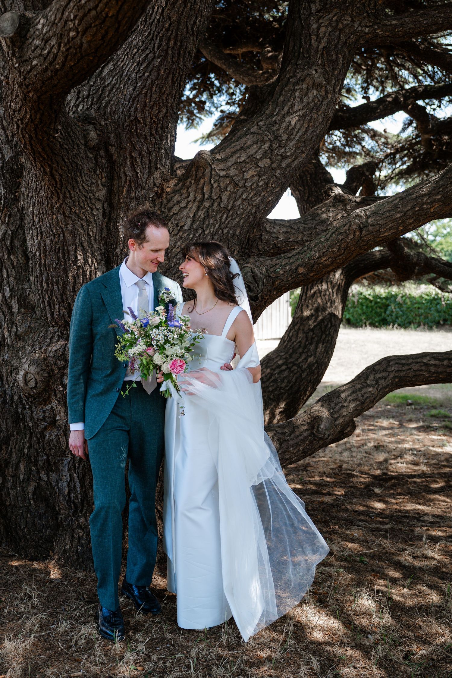 Bride and groom share a romantic moment under a large oak tree, bride holding a colorful bouquet in a white wedding gown.