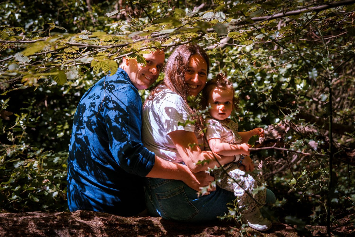 Family of three sitting together among dense green foliage, parents with young child exploring nature outdoors.