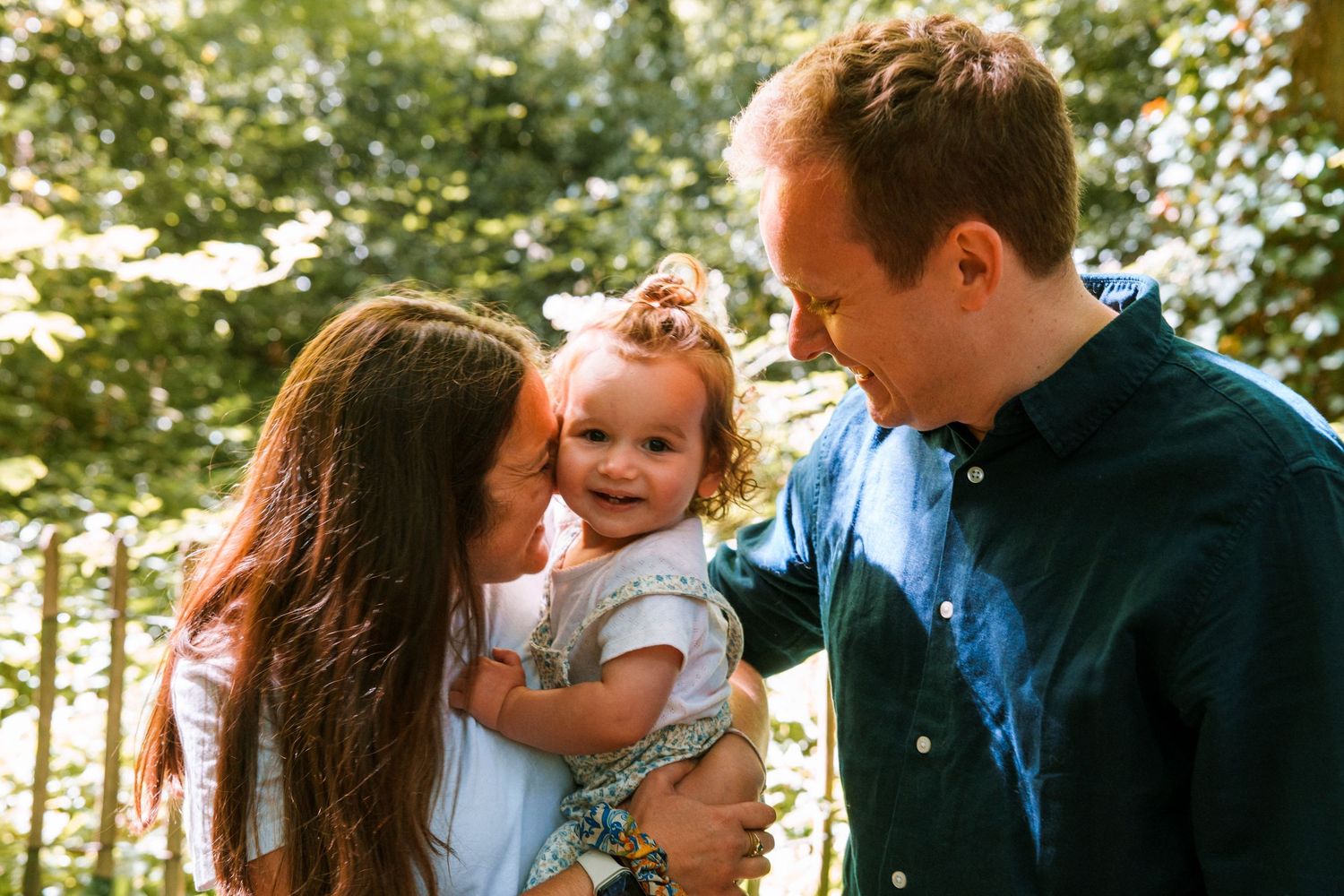 Happy family with mother kissing baby girl while father smiles outdoors in sunny green park setting.
