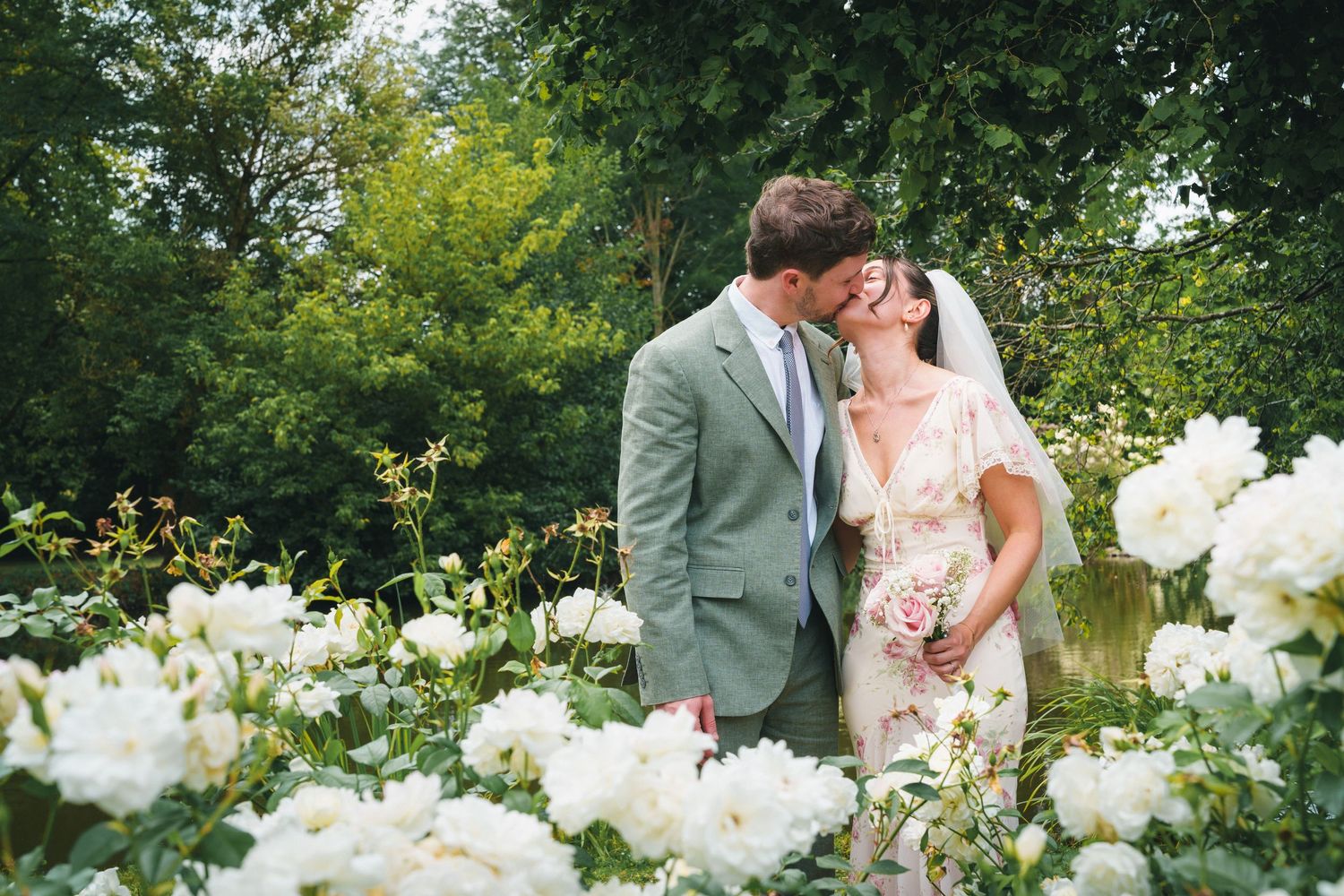 Bride and groom kissing romantically among white roses in lush garden after wedding ceremony.