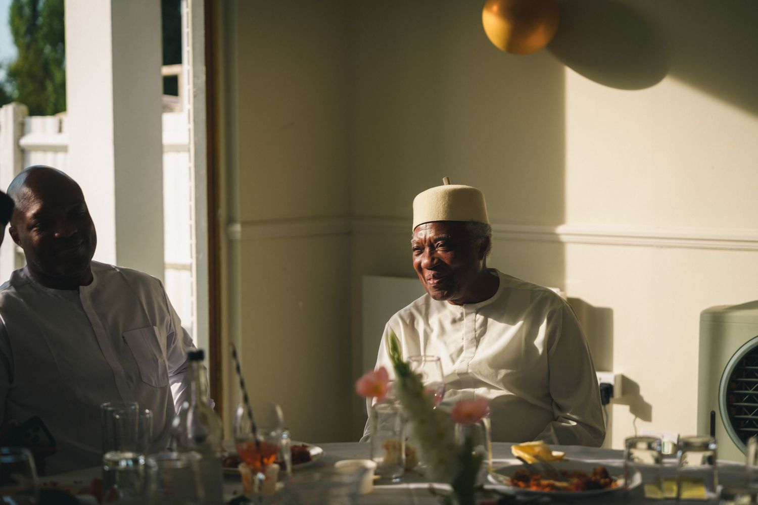 Two men in traditional white African attire sharing a meal at a table with food and drinks.