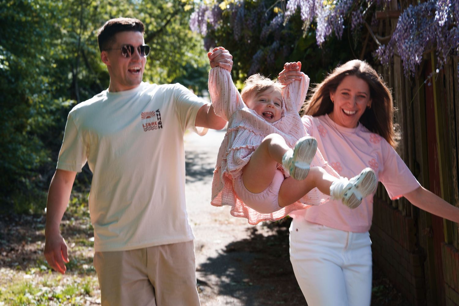 Happy family swinging toddler between parents outdoors in sunny garden with purple wisteria blooming in background.