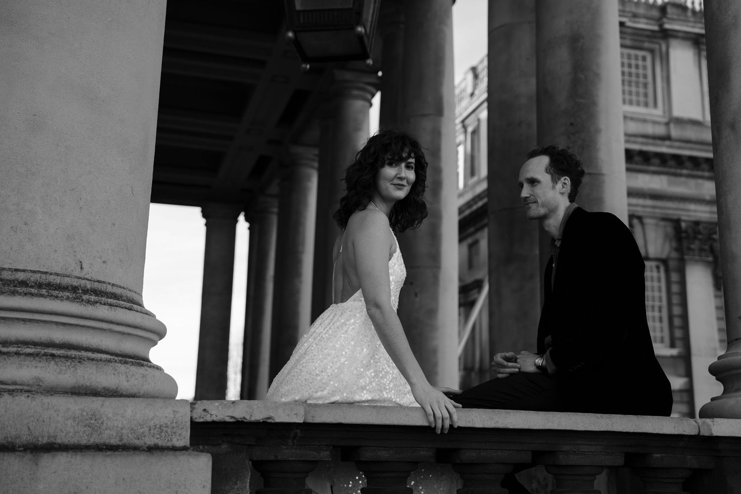 A romantic black and white portrait of a couple sharing a playful moment by classical architecture columns.