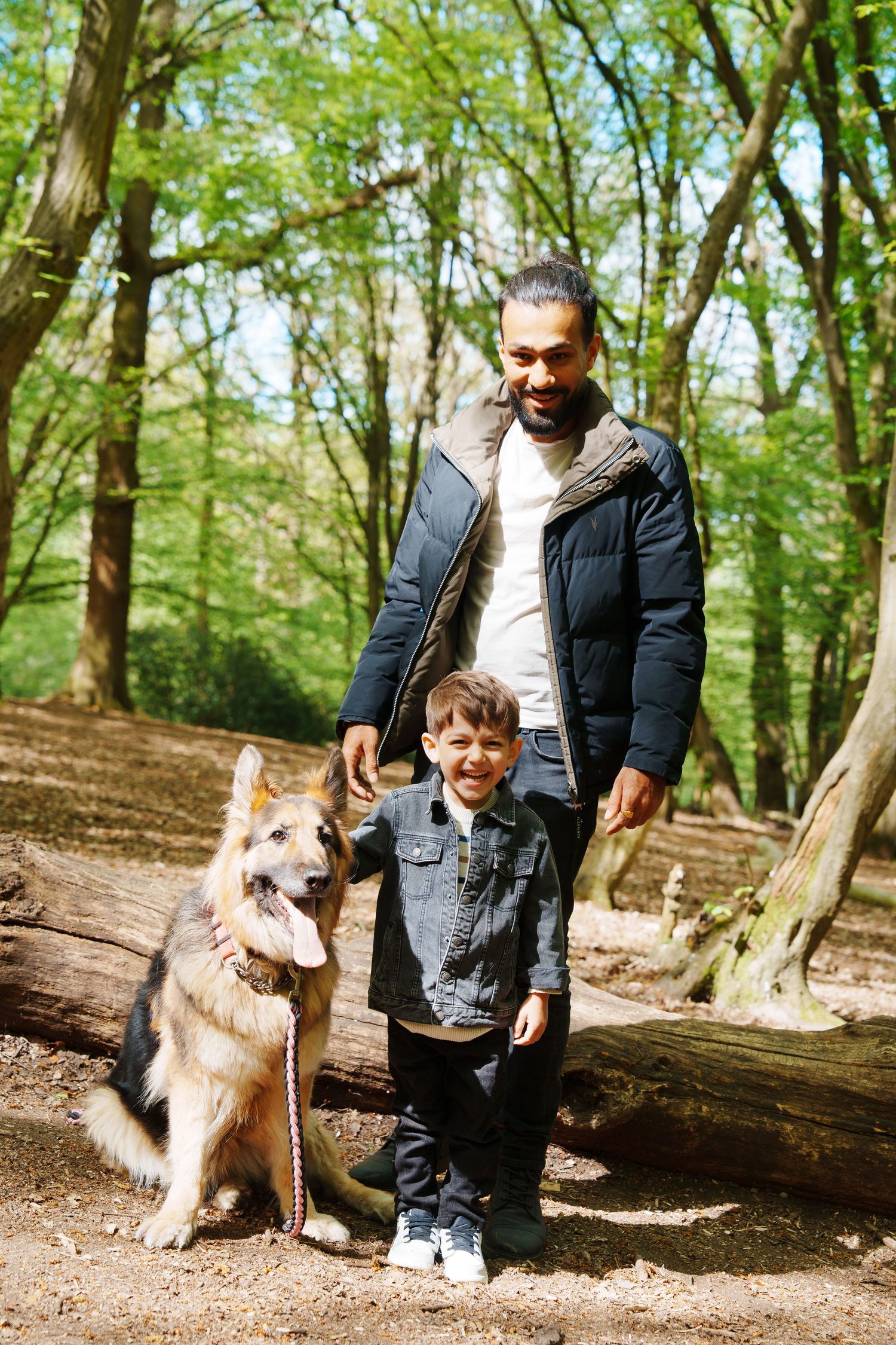 A man and young boy smile with a German Shepherd dog in a sunny woodland forest setting.