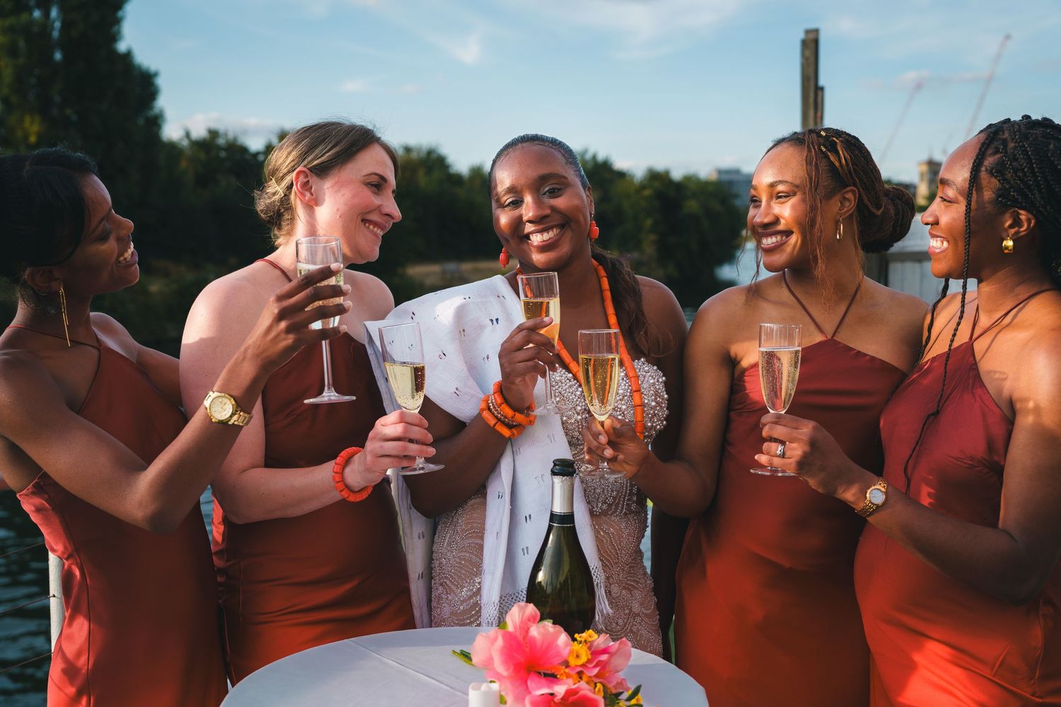 Bride in white suit toasting champagne with four bridesmaids in rust-red dresses at an outdoor waterfront celebration.