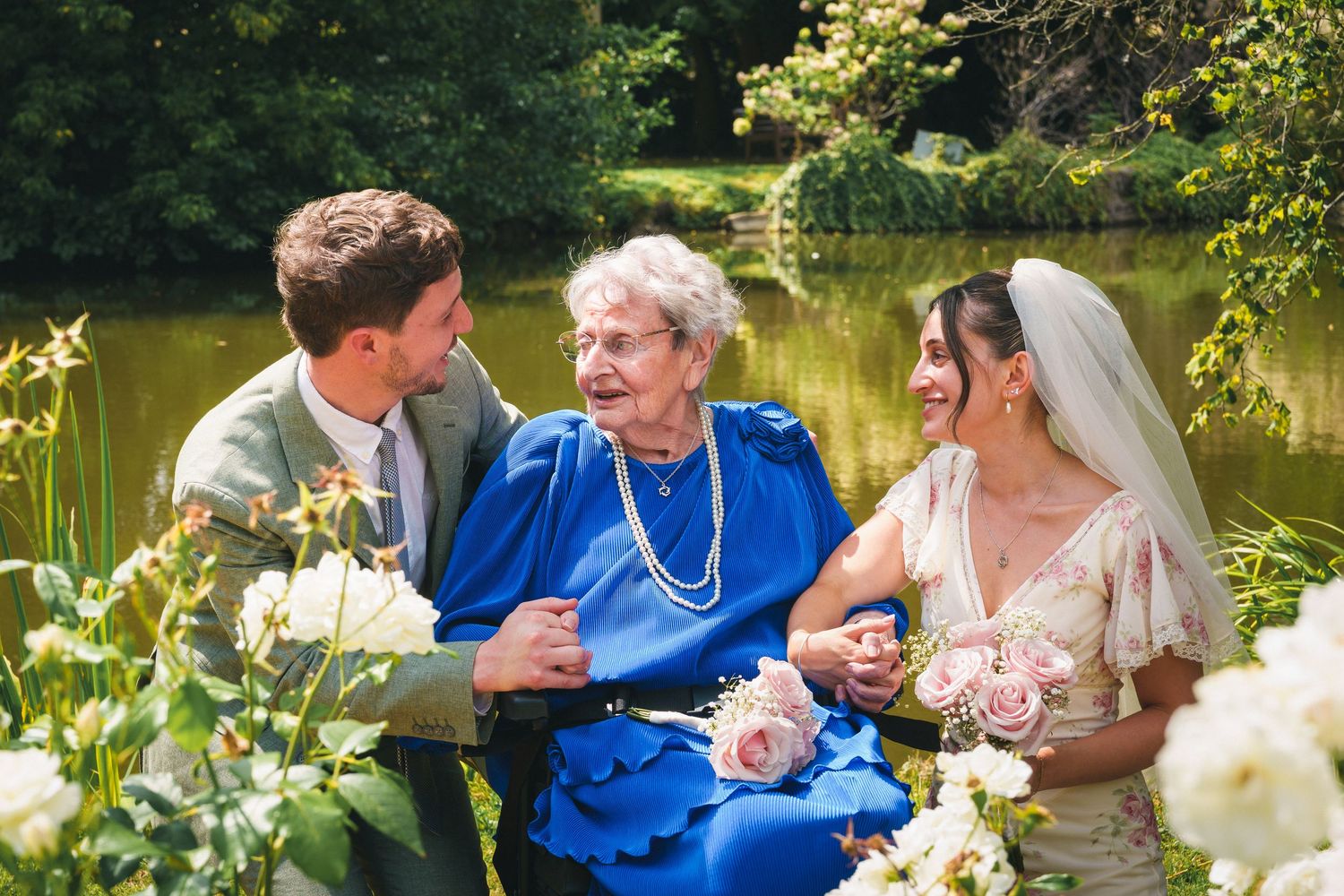 Bride and groom share a joyful moment with elderly woman in blue dress near a pond in a lush garden.