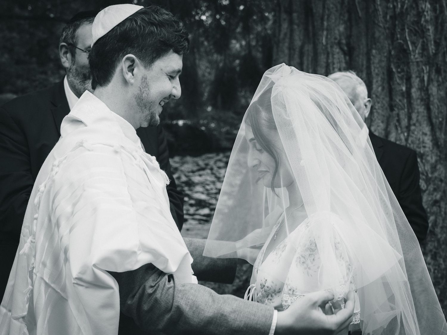 Black and white photo of a Jewish groom in tallit lifting bride's veil during outdoor wedding ceremony.