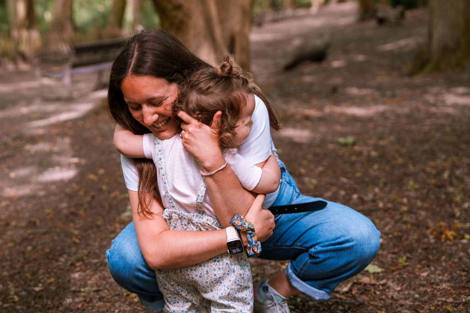 A mother crouching in a forest, lovingly embracing two young children in a warm, candid outdoor moment.