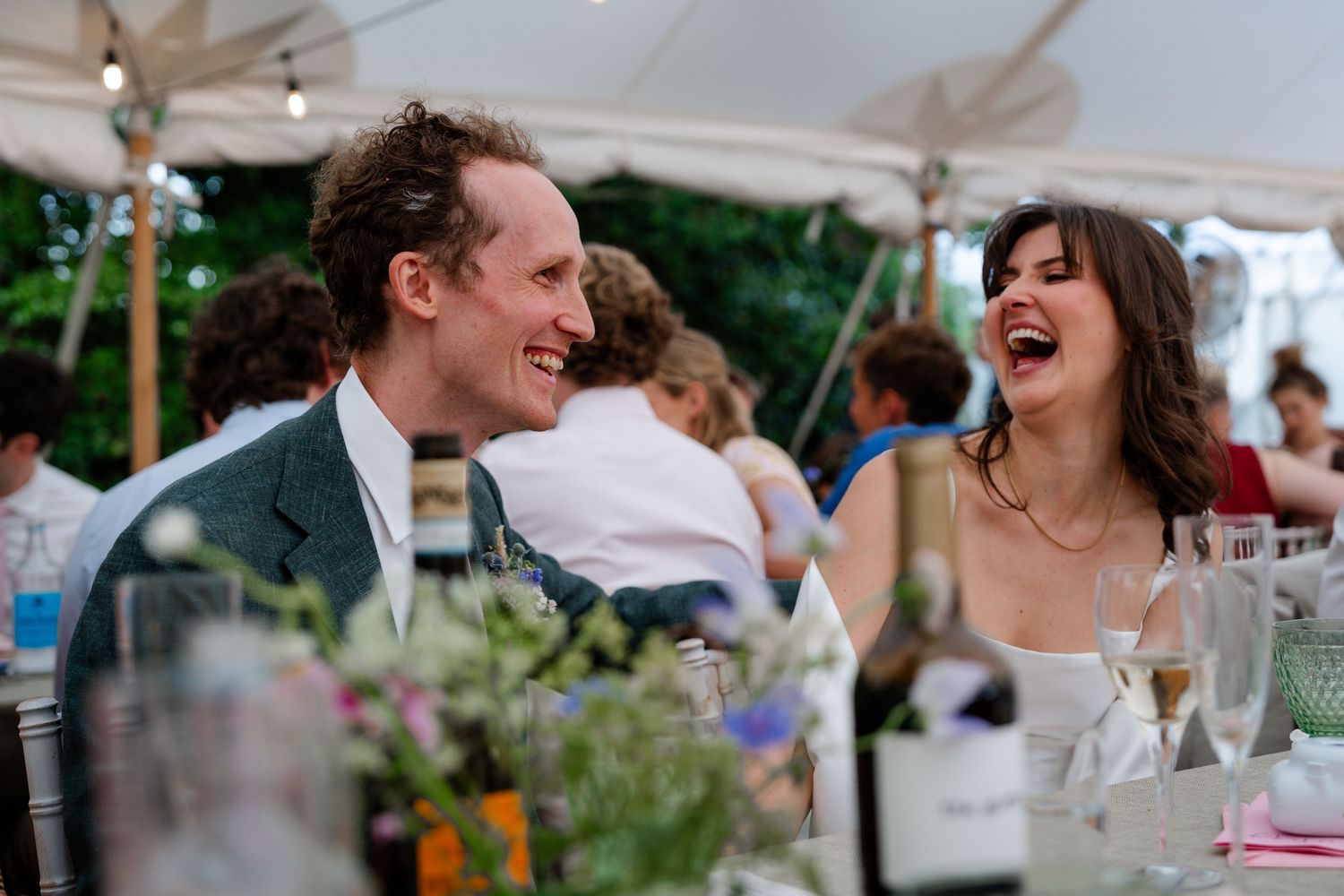 Bride and groom laughing joyfully at wedding reception dinner table under white tent.