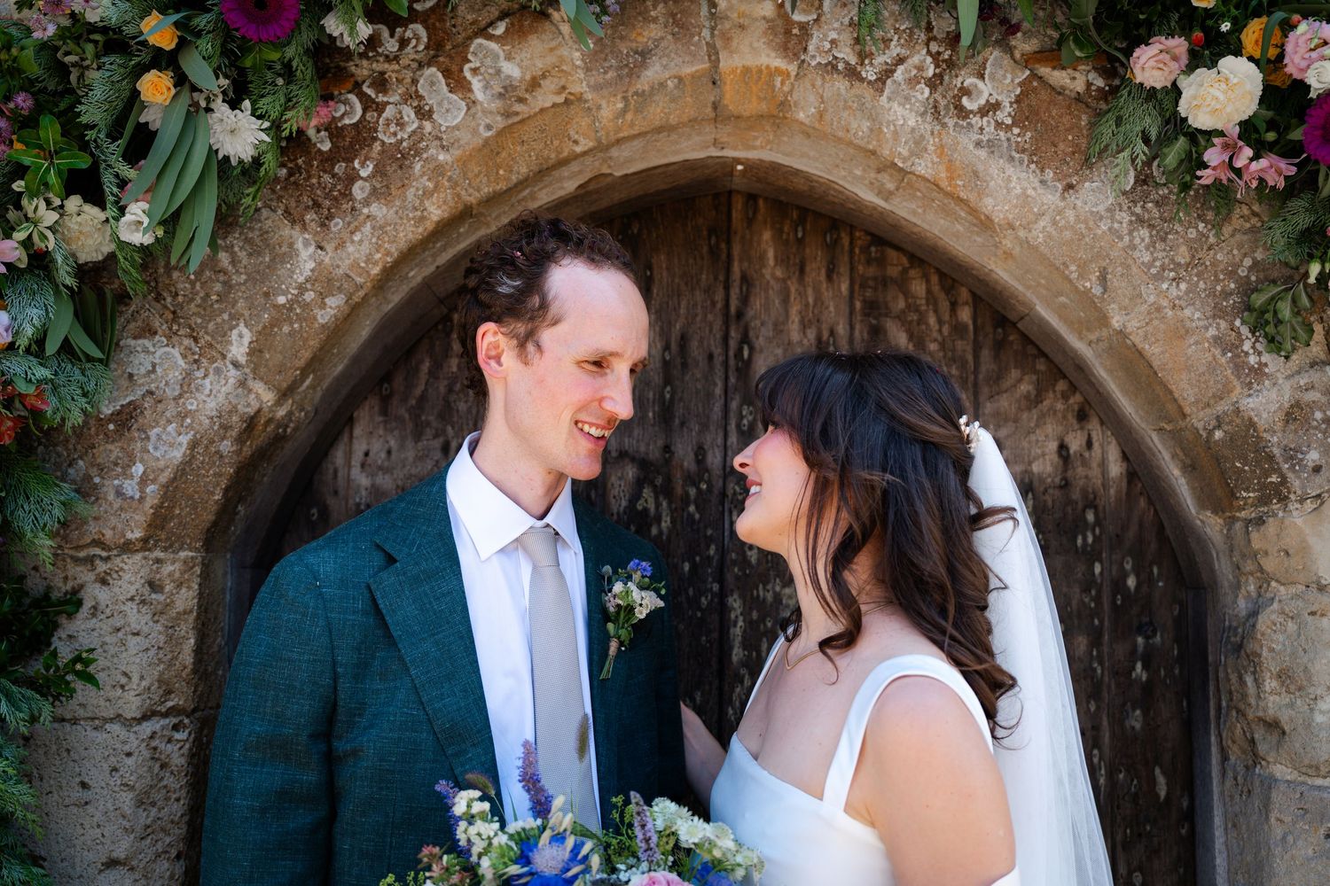 Bride and groom share a romantic moment in front of a stone church doorway adorned with colorful floral arrangements.