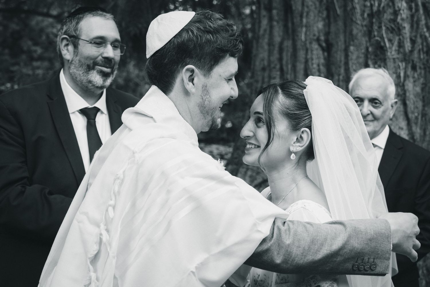 Black and white photo of a Jewish bride and groom embracing during their wedding ceremony under a chuppah.