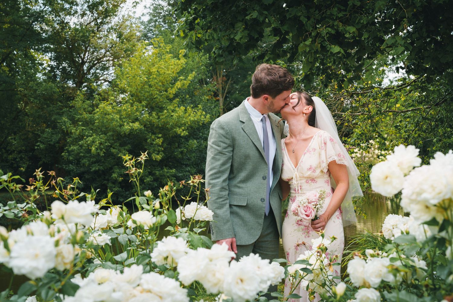 A bride and groom share a romantic kiss surrounded by lush white roses in a garden wedding setting.