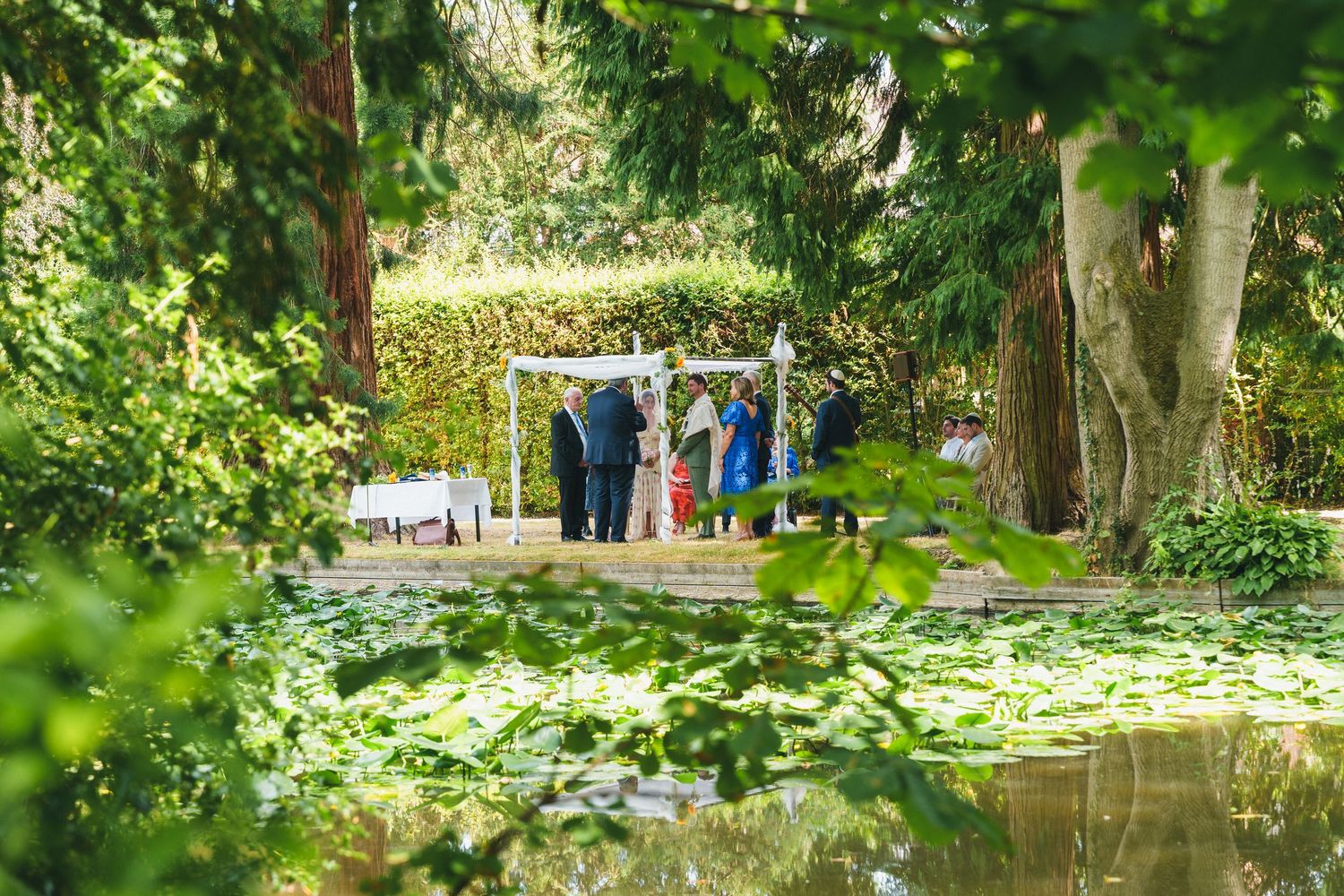 Outdoor Jewish wedding ceremony under chuppah beside tranquil garden pond surrounded by lush greenery.