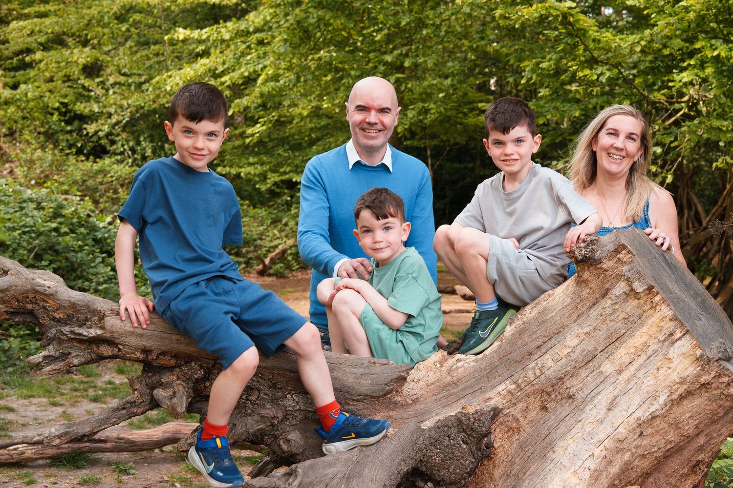 Family of five posing on fallen logs surrounded by lush green trees in an outdoor woodland setting.