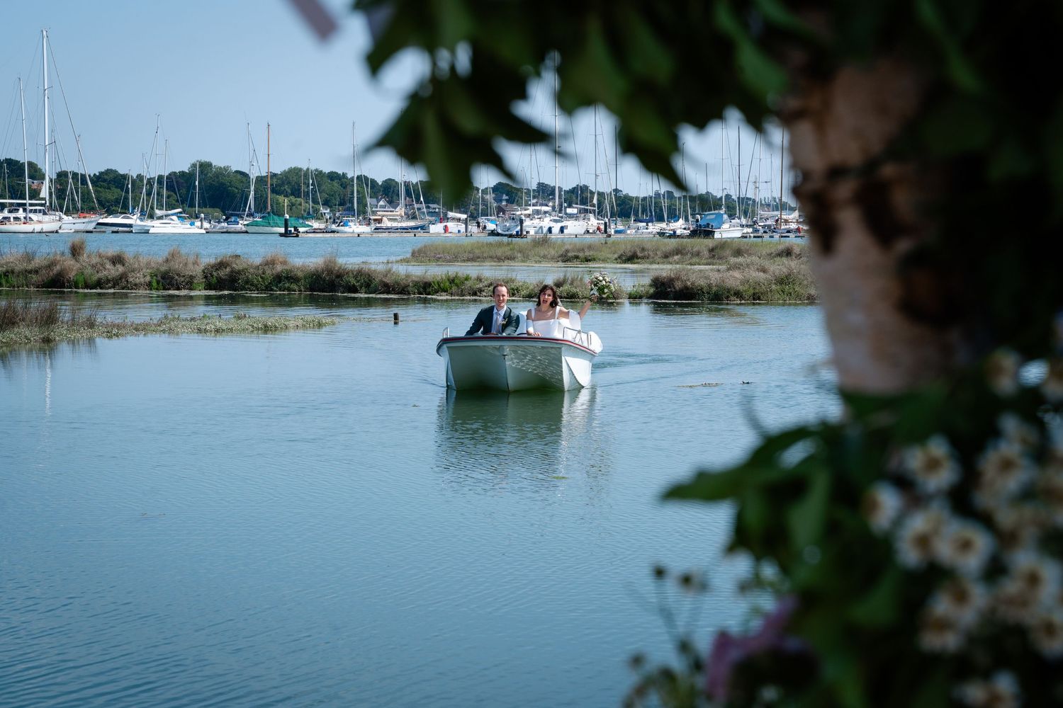 Couple arriving by small white boat on a calm marina waterway surrounded by sailboats and lush greenery.