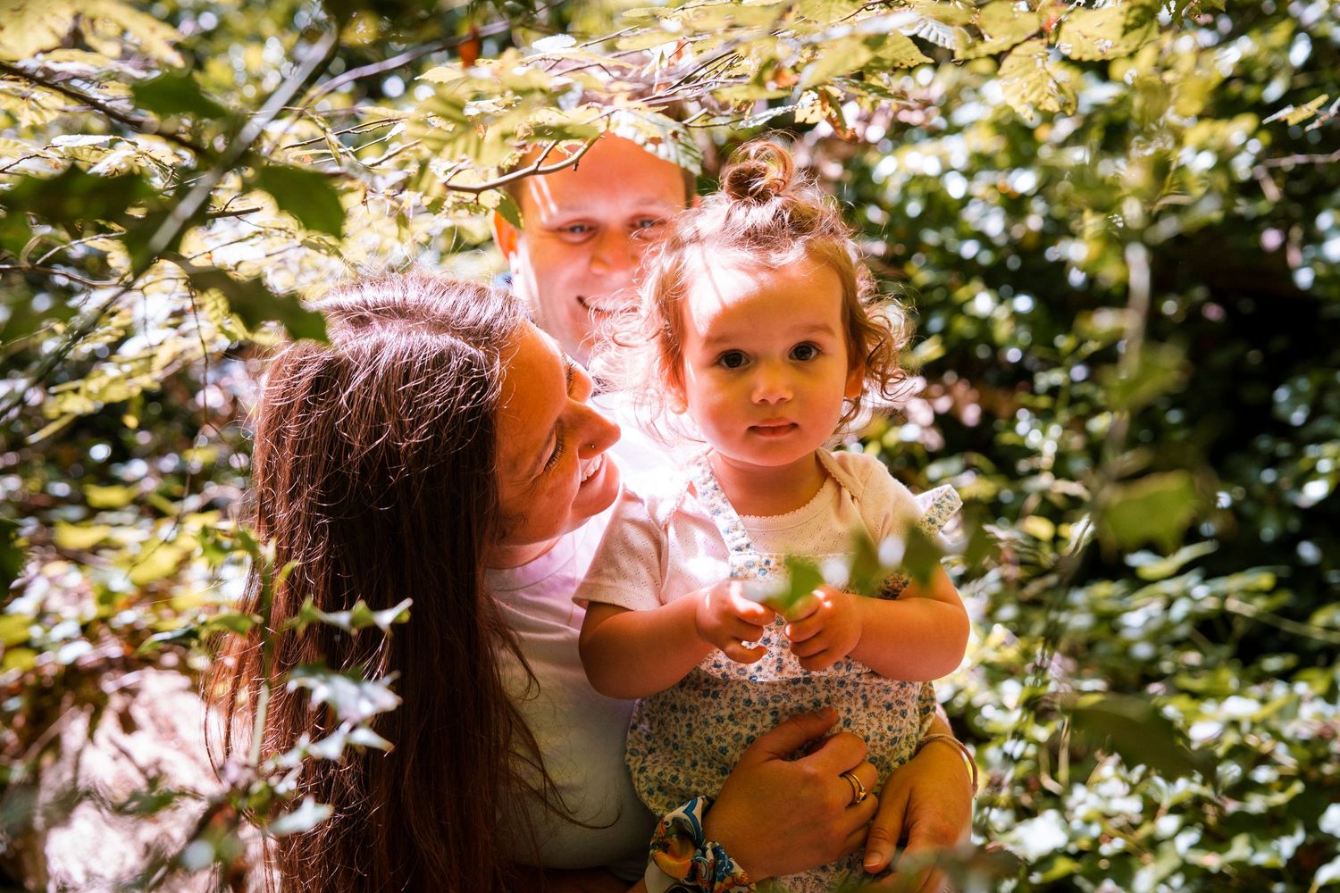 Family of three smiling among lush green leaves outdoors, parents holding young toddler girl in sunlit garden.