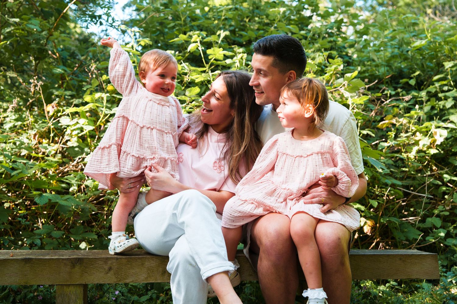 A happy family of four sits on a bench outdoors, wearing matching pink outfits, surrounded by lush green foliage.