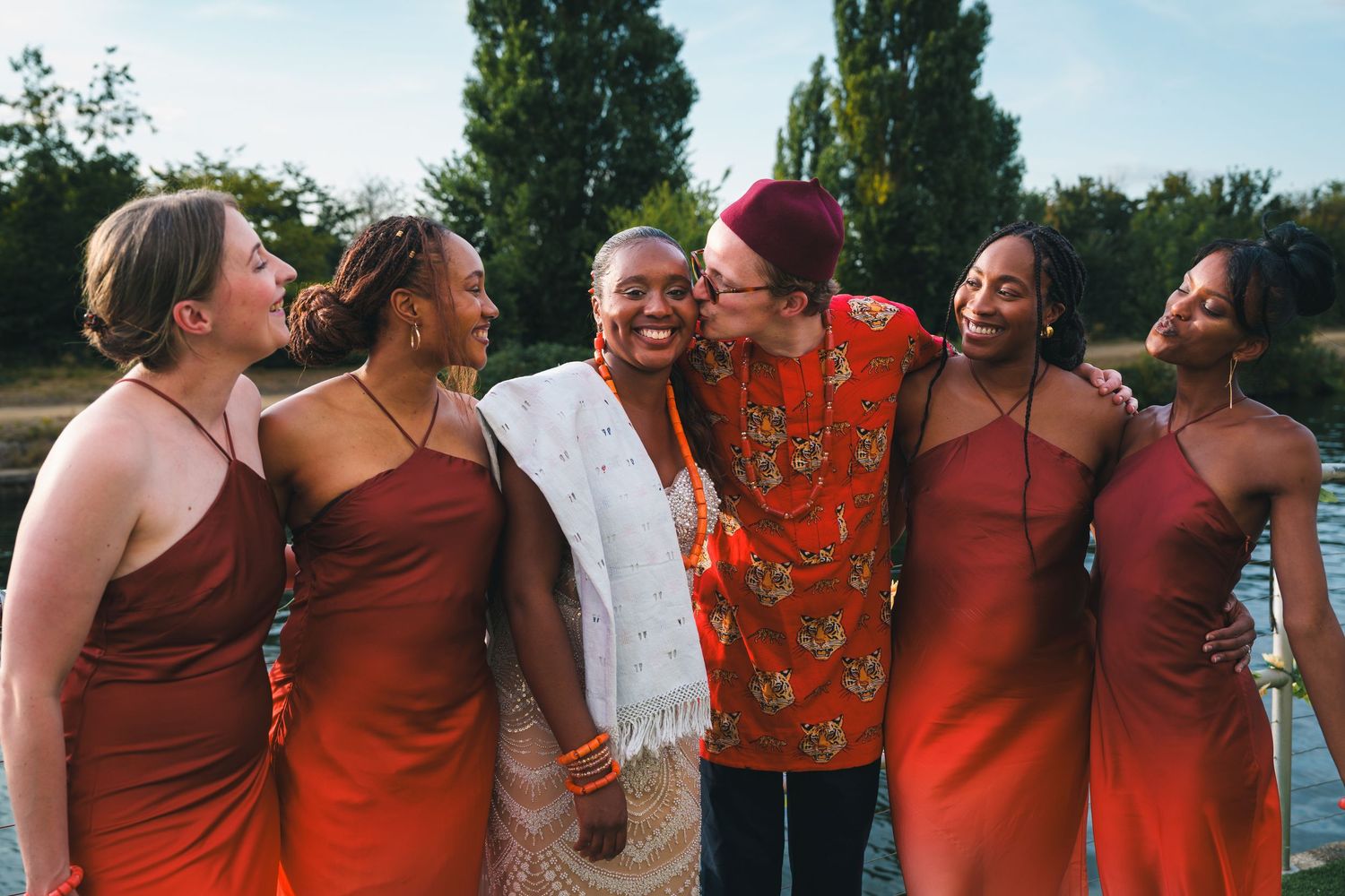 Group of women in red bridesmaid dresses laughing and celebrating outdoors near a lake.