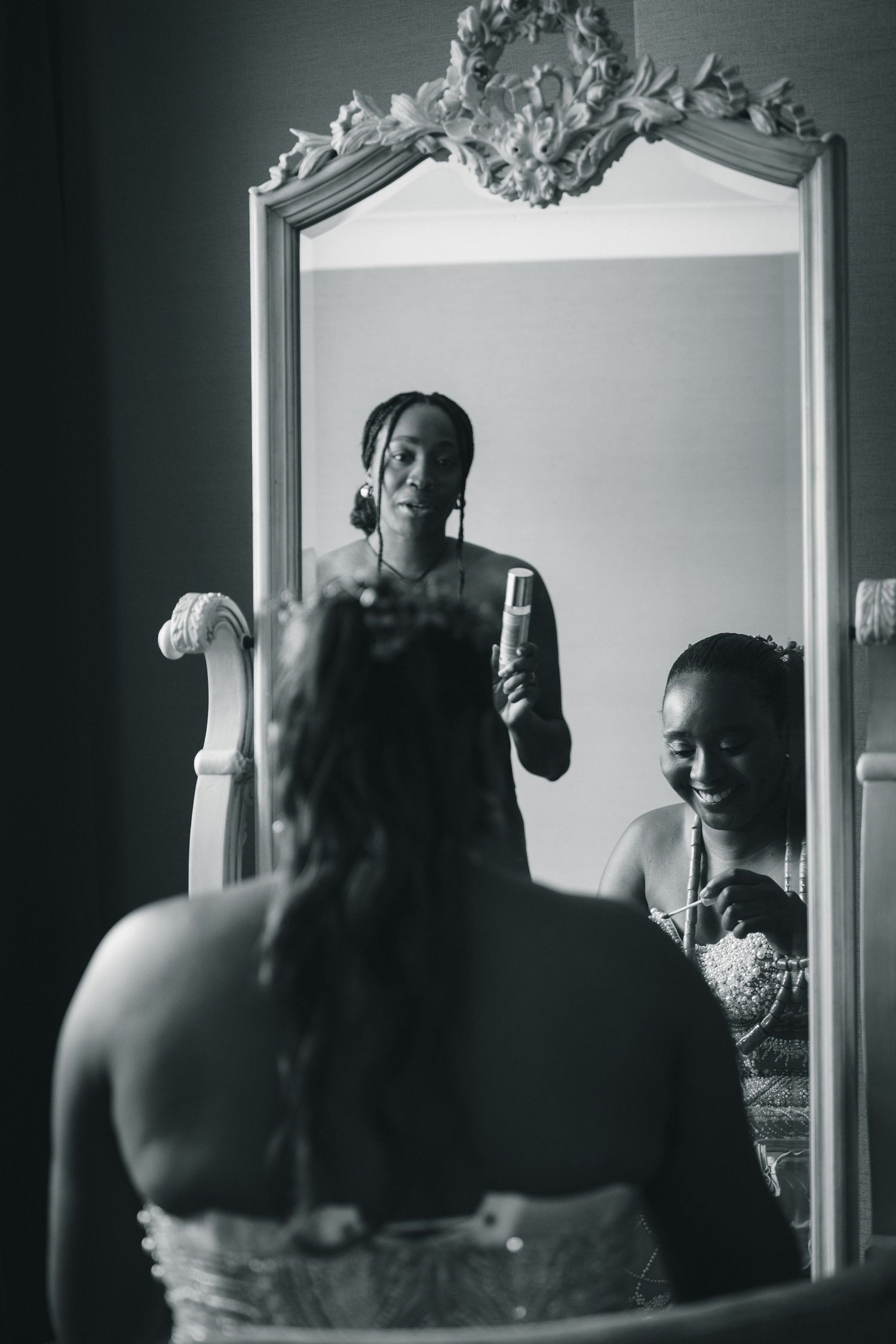 Black and white photo of a bride getting ready, reflected in an ornate mirror with a bridesmaid holding a phone.