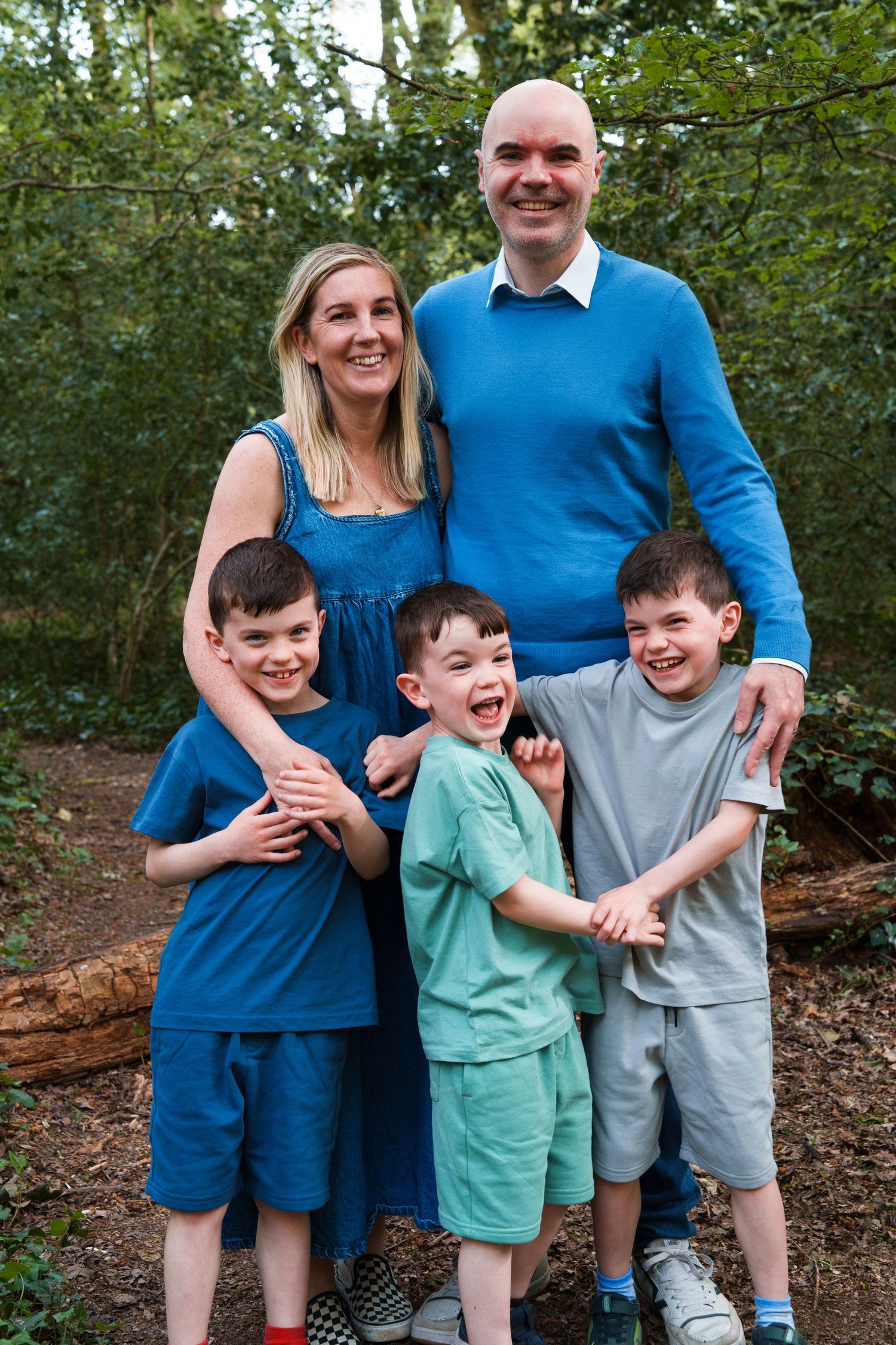 Happy family of five posing outdoors in blue outfits, two adults and three young boys smiling in a woodland setting.