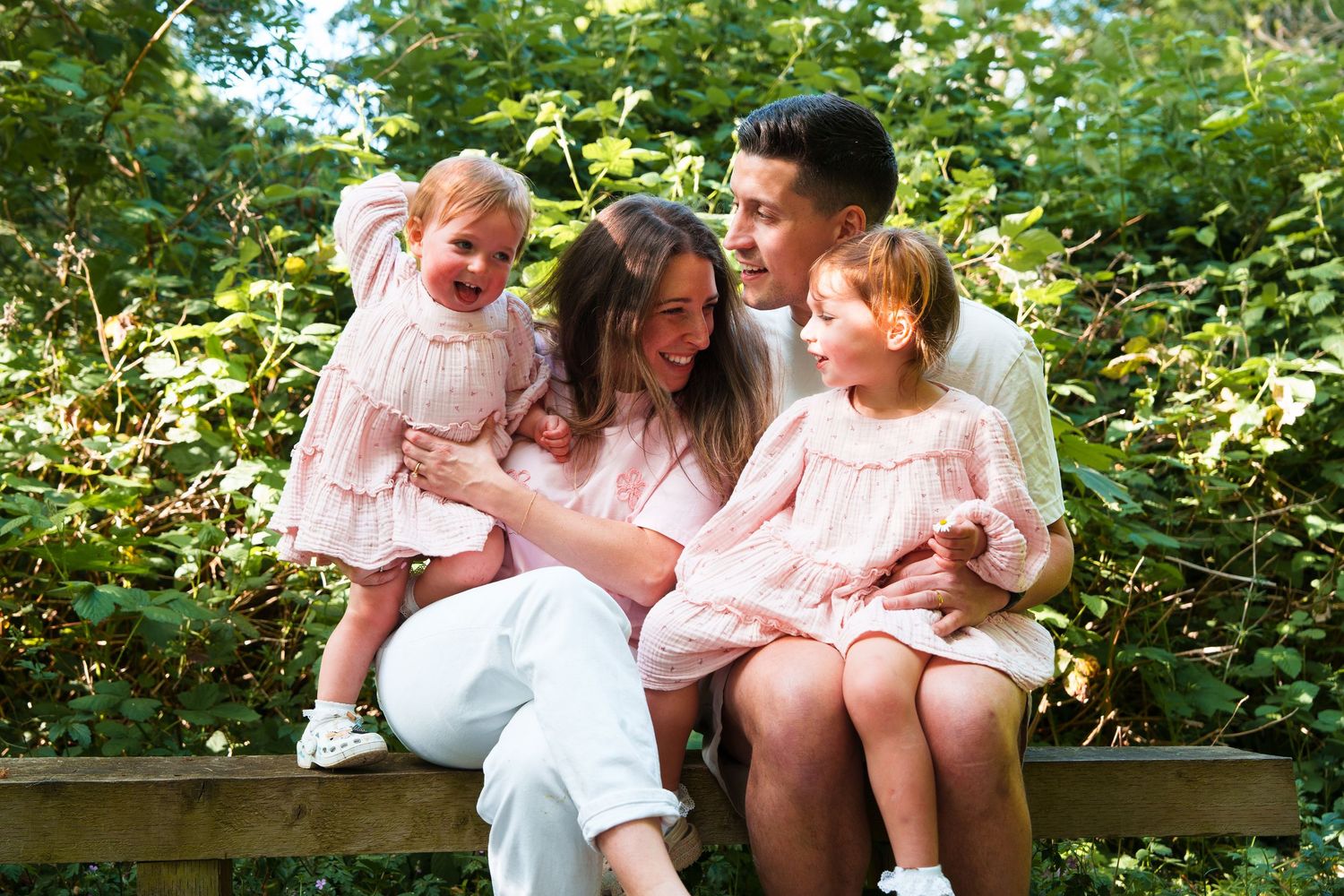 Happy family of four sitting on a wooden bench outdoors, two young girls in pink dresses with parents smiling.