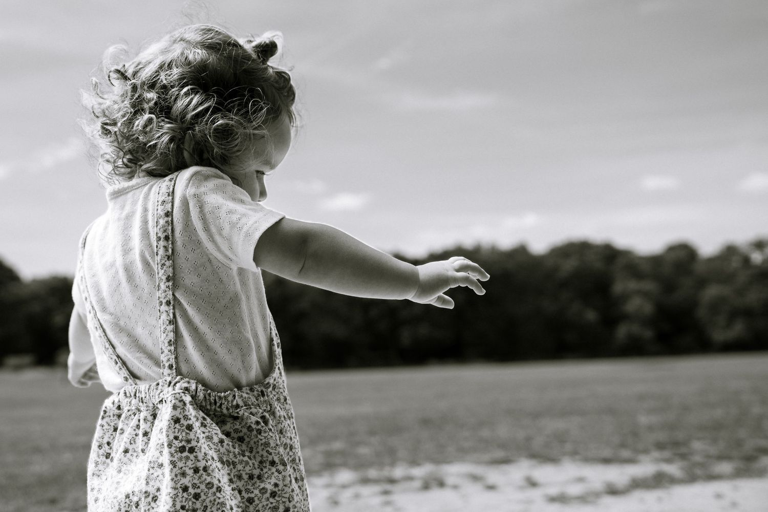 Black and white photo of a young curly-haired girl in a floral dress pointing toward an open field.
