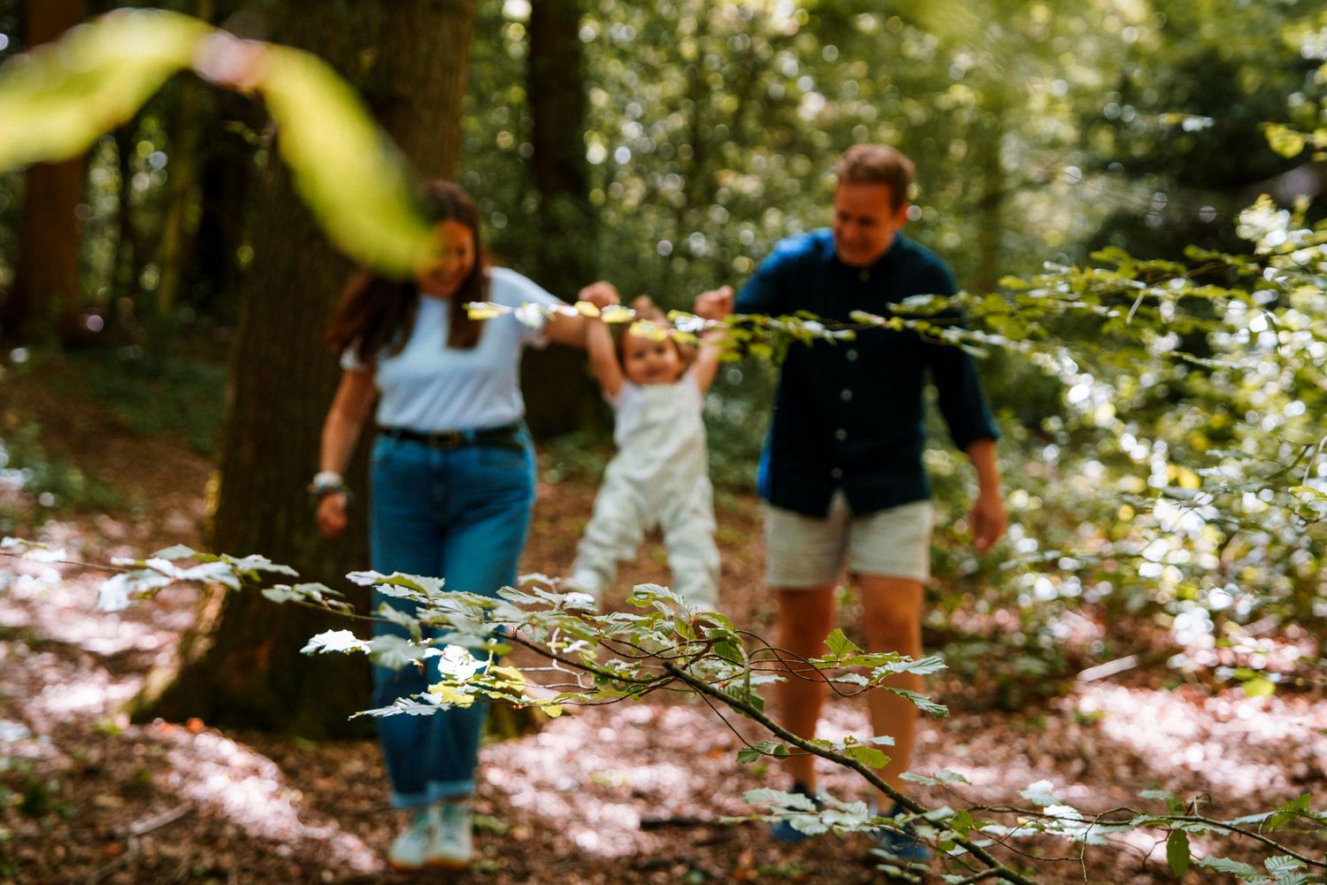 Family of three walking through a sunlit forest, parents holding toddler's hands among autumn leaves.